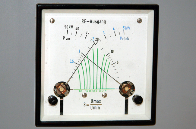 1) Kurzbeschreibung: Analoges Messinstrument mit Kreuzzeiger zur Anzeige des SWR mit Skalen für die vorlaufende und die rücklaufende Leistung und das Stehwellenverhältnis.

2) Ausführliche Beschreibung: Das Foto zeigt ein analoges Messinstrument mit Kreuzzeiger zur Anzeige des SWR. Links gibt es einen Zeiger für die vorlaufende Leistung („P_vor“) und rechts einen Zeiger für die rücklaufende Leistung („P_rück“). Unterhalb der beiden sich kreuzenden Skalen befindet sich eine horizontale Linie mit grünen Markierungen bei 1,3, 1,5, 1,6, 1,8, 2, 2,5, 3 und 4. Von jeder dieser Markierungen gehen grüne Linien nach oben, die im linken Teil nach links oben, im rechten Teil nach rechts oben geneigt sind. Die Ablesung erfolgt vom Kreuzungspunkt der beiden Zeiger aus entlang der jeweiligen grünen Linie zur Markierung auf der horizontalen Linie. Unten auf dem Messinstrument steht die Formel S = Umax / Umin.