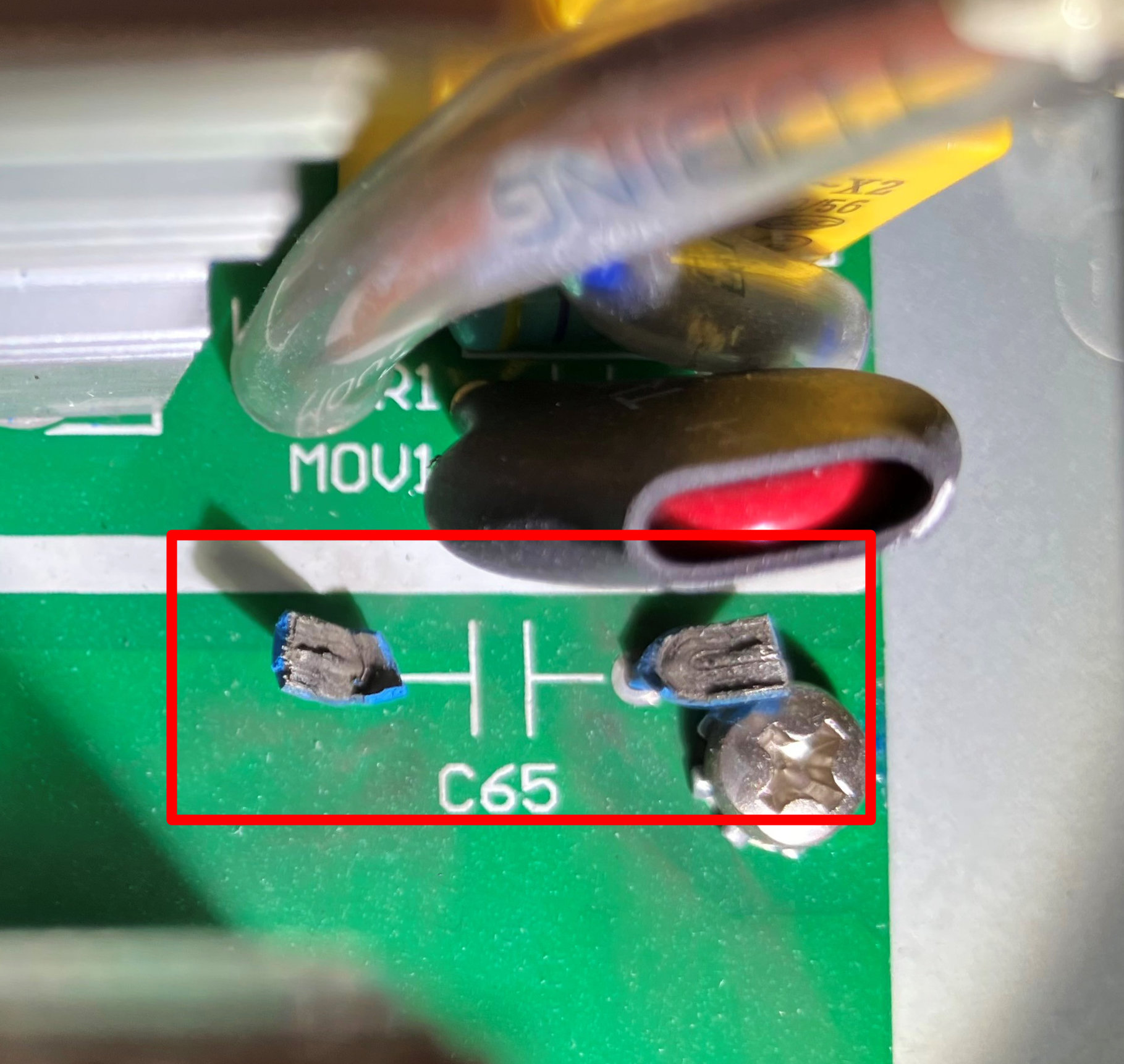 Dieser Alt-Text wurde noch nicht überprüft.

Zusammenfassung: Makroaufnahme einer grünen Leiterplatte, auf der der markierte Bestückungsplatz C65 mit zwei dunklen, beschädigt wirkenden Lötpads zu sehen ist, während oben drüber dicke Kabel verlaufen.

Detaillierte Beschreibung: In der Bildmitte ist ein roter Rahmen um den Bereich mit der weißen Aufschrift „C65“ und einem aufgedruckten Kondensatorsymbol gesetzt; die beiden zugehörigen Lötflächen sind uneben und dunkel verfärbt, mit Resten von Anschlussmaterial. Rechts daneben ist ein silberfarbener Schraubkopf (sternförfer Antrieb) zu sehen, der eine Ringkabelschuh-Lasche auf der Leiterbahn fixiert. Oberhalb des markierten Bereichs verlaufen zwei kräftige Kabel/Leitungen, eines dunkel mit rotem Innenmantel, das andere graumetallisch glänzend; sie werfen Schatten auf die Platine. Links ist der Rand eines kleinen Aluminiumkühlkörpers mit Rippen erkennbar. Auf der grünen Platine gibt es weiße Bestückungsdrucke, darunter teilweise lesbar „MOV1“, sowie eine breite weiße Leiterplattenmarkierung quer durch das Bild. Starke Punktbeleuchtung erzeugt Reflexe und harte Schatten.