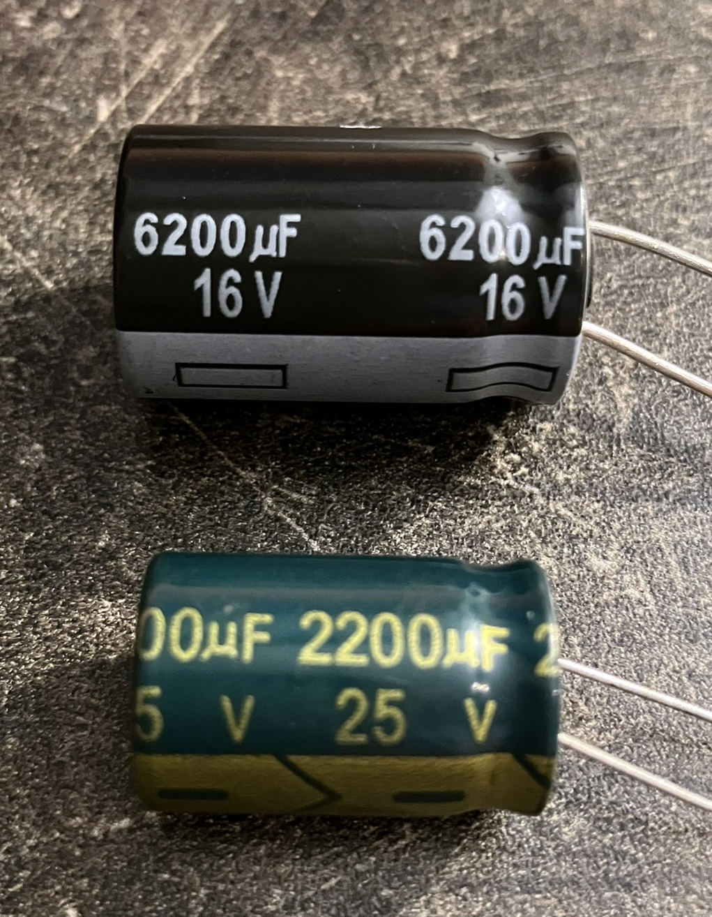 Dieser Alt-Text wurde noch nicht überprüft.

Kurzfassung: Zwei liegende Elektrolytkondensatoren mit Drahtanschlüssen auf einer dunklen, rauen Arbeitsfläche.

Detailbeschreibung: Oben ein größerer, zylindrischer Kondensator mit schwarzem Gehäuse und grauem Ring am unteren Ende; in weißer Schrift steht mehrfach „6200 µF 16 V“, die beiden Anschlussdrähte ragen nach rechts heraus. Darunter ein etwas kleinerer, dunkelgrüner Kondensator mit gelb-goldener Aufschrift „2200 µF 25 V“ und zusätzlichen Markierungen auf einem olivfarbenen Streifen; auch hier führen zwei Drähte nach rechts. Die Oberfläche zeigt Kratzer und Staub, das Licht erzeugt glänzende Reflexe auf den Kondensatoren, die nahezu parallel zueinander liegen.