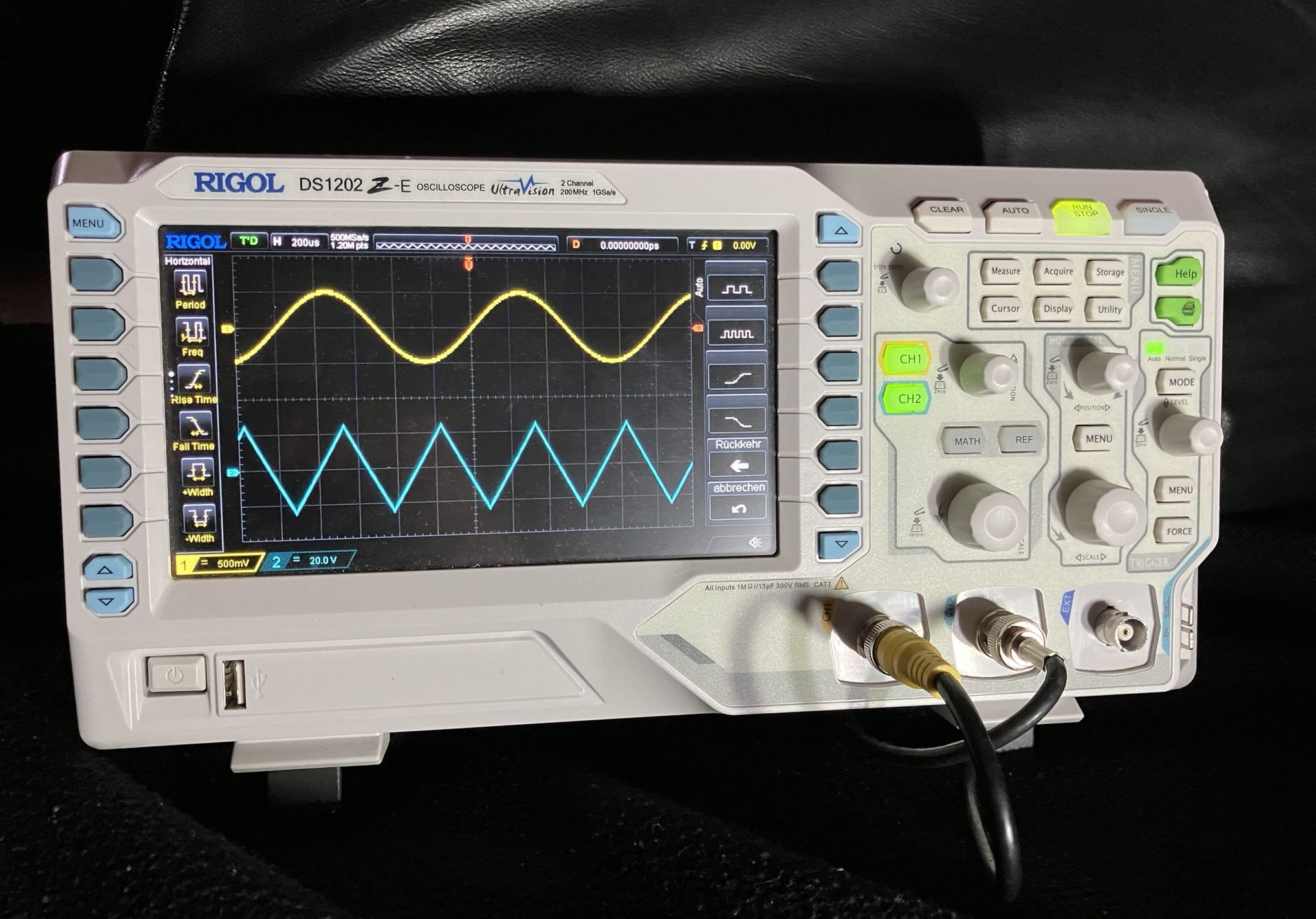 Dieser Alt-Text wurde noch nicht überprüft.

1) Zusammenfassung: Ein digitales Rigol-Oszilloskop zeigt gleichzeitig eine gelbe Sinuskurve und eine blaue Dreieckskurve auf seinem Display.

2) Detaillierte Beschreibung: Das Gerät ist ein weißes Tischoszilloskop mit der Aufschrift „RIGOL DS1202Z‑E“ und „UltraVision“, fotografiert auf dunklem Hintergrund. Auf dem farbigen Bildschirm ist ein schwarzgraues Raster mit zwei Messkurven zu sehen: oben eine gelbe, weich geschwungene Sinuslinie, darunter eine blaue, gleichmäßig gezackte Dreieckslinie; links und rechts vom Messfeld stehen Symbolleisten mit gelben Funktionspiktogrammen (z. B. Periodendauer, Frequenz) und rechts vertikale Softkeys mit Kurven-Symbolen sowie deutschen Beschriftungen wie „Rückkehr“ und „abbrechen“. Am unteren Displayrand sind farbige Kanalbalken und Skalierungsangaben erkennbar. Rechts neben dem Display befinden sich zahlreiche Tasten und Drehregler; die Kanal-Tasten „CH1“ und „CH2“ leuchten grün. Unten rechts sind zwei BNC‑Buchsen mit einem gelben und einem schwarzen Koaxkabel belegt, eine weitere Buchse daneben ist frei. Links unten am Gerät sind ein Netzschalter und ein USB‑Anschluss sichtbar.