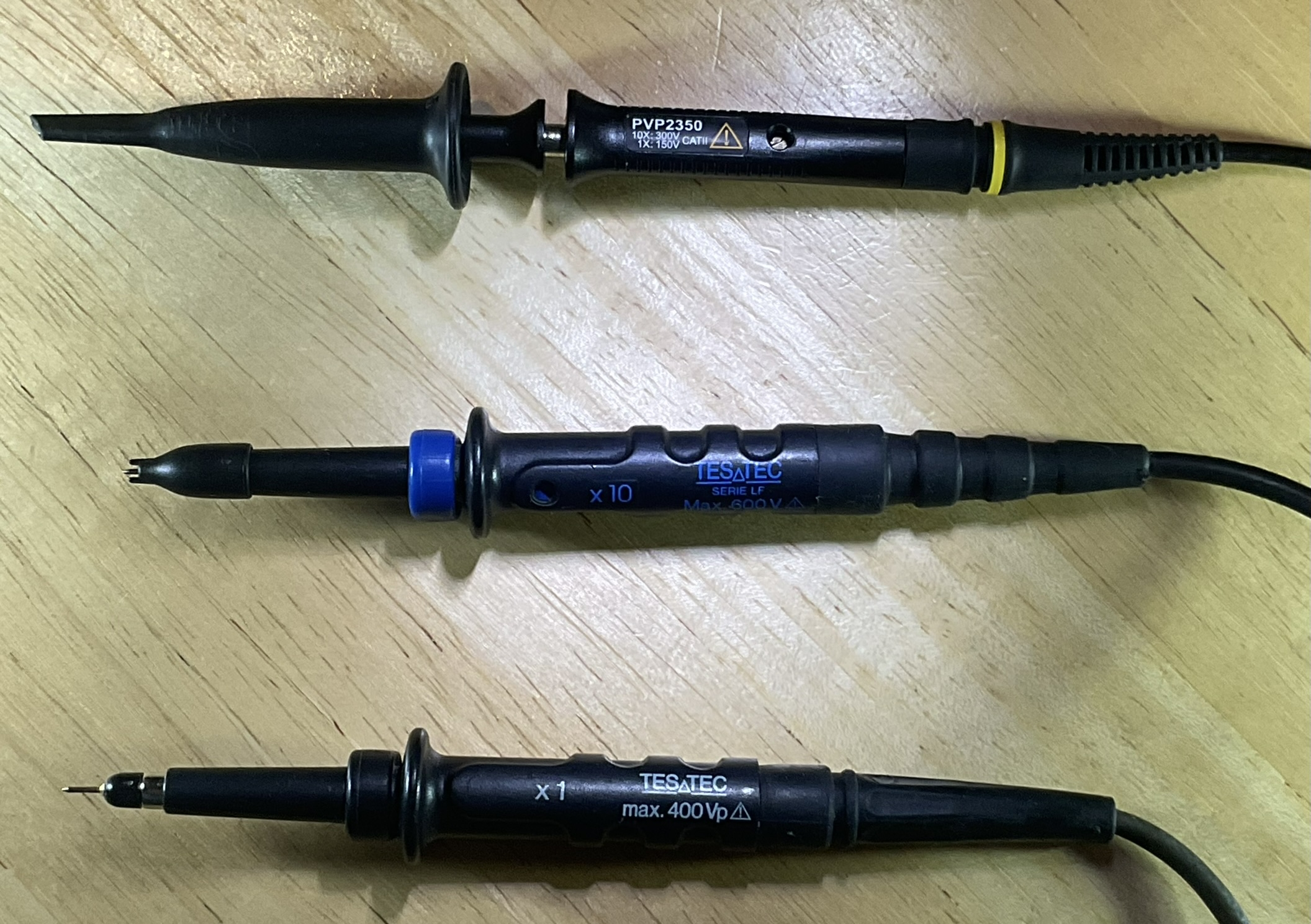 Dieser Alt-Text wurde noch nicht überprüft.

1) Kurzzusammenfassung: Drei schwarze Oszilloskop‑Tastköpfe liegen parallel auf einer hellen Holzoberfläche.

2) Detailbeschreibung: Die drei Sonden sind horizontal ausgerichtet, mit den Spitzen nach links und den Kabeln nach rechts. Oben befindet sich ein schwarzer Tastkopf mit der Aufschrift „PVP2350“, Warnsymbol und gelbem Ring an der Zugentlastung; die Spitze ist mit einer länglichen schwarzen Schutzkappe versehen. In der Mitte liegt ein schwarzer Tastkopf mit blauem Einstellring und der blauen Beschriftung „x10“, „TESATEC“, „SERIE LF“, „Max. 600V“; an der Spitze ist eine kleine Haken- oder Steckspitze sichtbar, teils von einer kurzen Kappe umgeben. Unten liegt ein weiterer schwarzer Tastkopf mit weiß/blauer Beschriftung „x1“, „TESATEC“, „max. 400Vp“; hier ist eine feine, nadelförmige Metallspitze freiliegt. Die Gehäuse haben gerippte Griffflächen, die Kabel führen nach rechts aus dem Bild, und die Holzoberfläche zeigt deutliche Maserung.