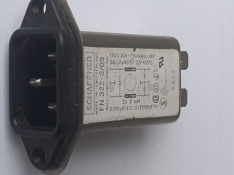 Dieser Alt-Text wurde noch nicht überprüft.

1) Kurzzusammenfassung: Ein flanschmontierbarer IEC-Netzeinbaustecker mit integriertem Filter von Schaffner liegt auf weißem Hintergrund.

2) Detaillierte Beschreibung: Links ist die schwarze Kunststofffront mit einem dreipoligen IEC-C14-Einlass und zwei Befestigungsbohrungen zu sehen, daran schließt sich ein längliches Metallgehäuse an. Auf der Oberseite klebt ein weißes Typenschild mit der Aufschrift „SCHAFFNER – MADE IN SWITZERLAND – FN 322-…“, mit Angaben wie „110/250 V~ 50/60 Hz“ und „3 A“, einem kleinen Schaltbild (mit Beschriftungen „2× 2 mH“, „0,015 µF (X2)“ und „2× 2200 pF (Y)“) sowie mehreren Prüf- und Zulassungszeichen. Rechts ragen zwei Flachsteckkontakte aus dem Gehäuse heraus; an der Seite ist eine Zahl (z. B. „8402“) aufgedruckt. Die Aufnahme ist leicht seitlich gedreht, die Farben sind Schwarz, Grau und Weiß, mit leichten Gebrauchsspuren am Gehäuse.