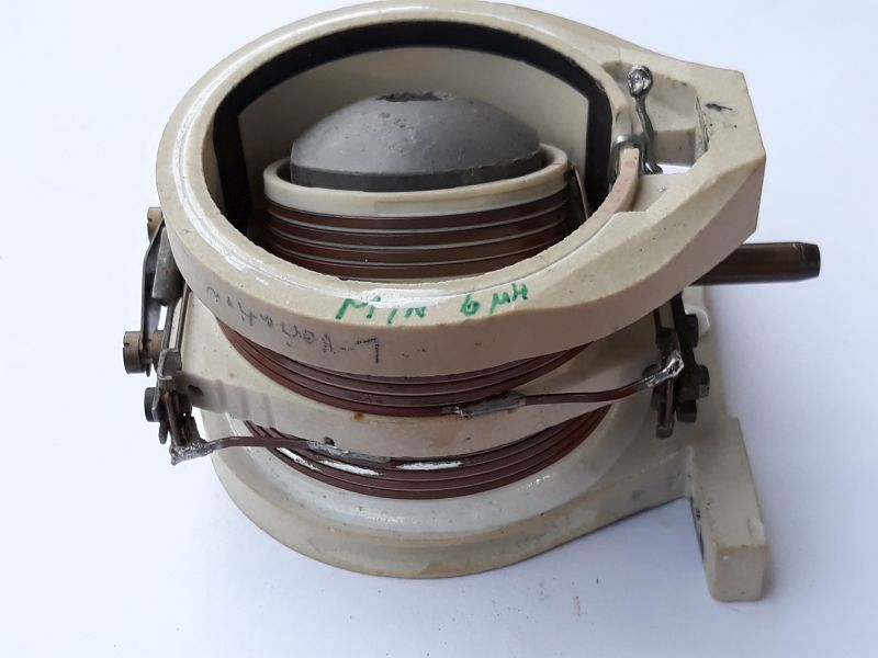 Dieser Alt-Text wurde noch nicht überprüft.

1. Zusammenfassung: Nahaufnahme eines cremefarbenen Spulengehäuses mit braunem Kupferdraht, Metallkontakten und einer grauen Kuppel im Inneren, auf weißem Hintergrund.

2. Detaillierte Beschreibung: Das Objekt besteht aus einem ringförmigen, hellen Gehäuse mit mehreren Lagen braun lackiertem Kupferdraht, der um einen keramischen Träger gewickelt ist. Im Zentrum sitzt eine graue, kuppelförmige Innenteile. Auf dem oberen Gehäuserand steht handschriftlich in grüner Farbe „MIN 6 mH“. An den Seiten sind Metallklammern, Schrauben und Lötfahnen zu sehen; ein dünner Metallkontakt verläuft entlang der Wicklung. Rechts ragt ein kurzer, zylindrischer Stift aus dem Gehäuse, links befinden sich einstellbare Schraubelemente. Das Gerät steht auf einer zweiteiligen Fußbasis mit Befestigungslöchern. Oberflächen zeigen Kratzer, Kleberreste und Gebrauchsspuren; der Hintergrund ist schlicht weiß.