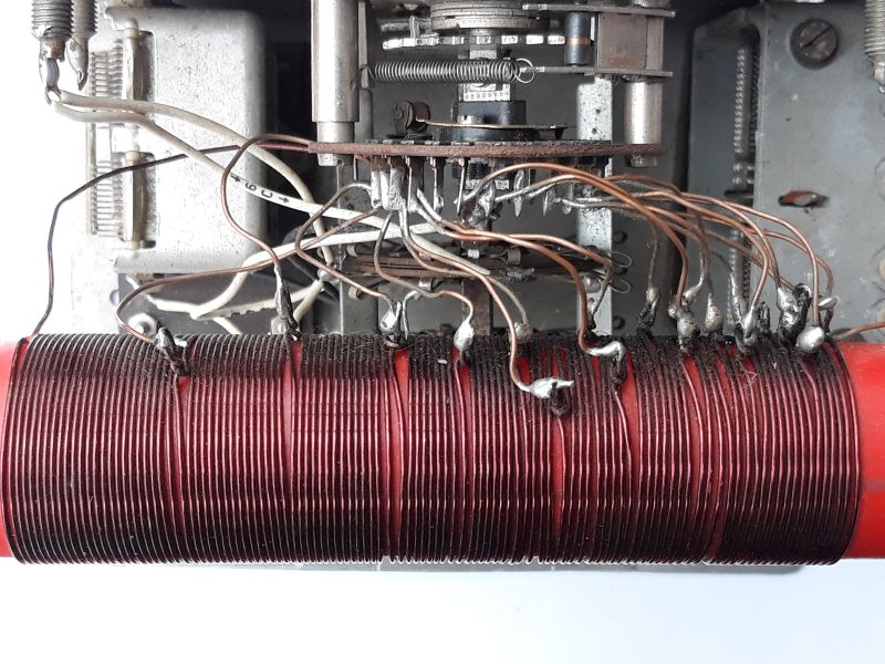 Dieser Alt-Text wurde noch nicht überprüft.

Zusammenfassung: Nahaufnahme des Innenraums eines elektronischen Geräts mit einer dicht gewickelten roten Zylinderspule und zahlreichen Drahtabgriffen, die zu einem mehrstufigen Drehschalter im Metallchassis führen.

Detaillierte Beschreibung: Im unteren Bildbereich liegt horizontal eine rote, zylindrische Spule, eng mit dunkelrotem Kupferdraht bewickelt. Entlang der oberen Spulenseite sind viele Abgriffe sichtbar: dünne, bräunliche lackierte Drähte, die an einzelnen Windungen angelötet sind; an den Lötstellen sitzen silbrig glänzende Lottropfen, teils mit dunklen Verfärbungen an der Umgebung. Die Abgriffe verlaufen bogenförmig nach oben und vorne zu einem gestapelten, kreisförmigen Drehschalter mit mehreren Isolatorscheiben und radial angeordneten Lötfahnen. In der Schaltermitte sitzt eine Metallwelle mit Federn und kleinen mechanischen Teilen. Links und rechts erkennt man das graue Metallchassis mit Schrauben, Federn und Halterungen; einzelne weiße und graue Leitungen führen in den Aufbau. Der Hintergrund ist überwiegend grau-metallisch, unter der Spule ist ein heller, weißer Untergrund zu sehen. Der Gesamteindruck ist technisch, gebraucht und funktional, mit leicht unordentlich geführten Drähten.