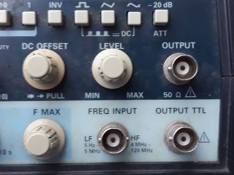 Dieser Alt-Text wurde noch nicht überprüft.

Zusammenfassung: Ausschnitt der Frontplatte eines elektronischen Messgeräts mit mehreren Drehknöpfen und BNC-Buchsen für Ausgang und Frequenzeingang.

Detaillierte Beschreibung: Oben verläuft eine Reihe hellgrauer, quadratischer Tasten mit Symbolen (u. a. Wellenformen) und Beschriftungen wie INV, DC und -20 dB (ATT). Darunter links ein großer, beigefarbener Drehknopf mit der Beschriftung DC OFFSET und dem Hinweis → PULL; in der Mitte ein gleich großer Knopf LEVEL mit Skala von MIN bis MAX. Rechts daneben eine silberne, runde BNC-Buchse mit der Beschriftung OUTPUT und 50 Ω sowie einem Warnsymbol. Im unteren, hellblauen Bereich befindet sich links ein weiterer Drehknopf F MAX. In der Mitte ist eine BNC-Buchse unter der Überschrift FREQ INPUT, umgeben von den Angaben LF 5 Hz–5 MHz und HF 4 MHz–120 MHz. Rechts unten eine weitere BNC-Buchse mit der Beschriftung OUTPUT TTL und einem Warnsymbol. Die Front ist dunkelblau mit hellblauen Feldern; die Bedienelemente wirken leicht benutzt.