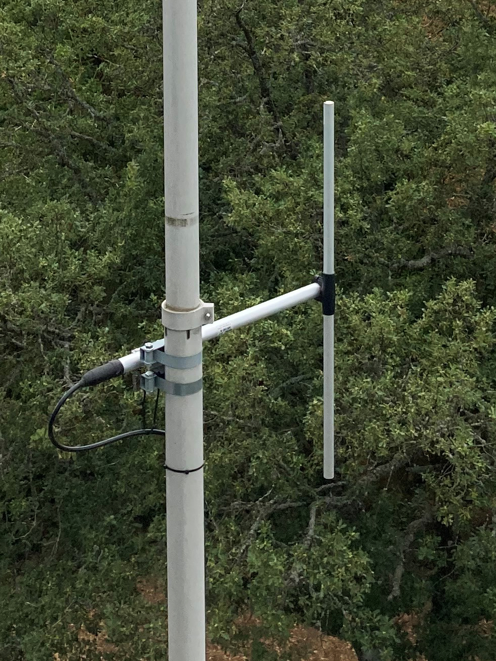 Dieser Alt-Text wurde noch nicht überprüft.

1) Kurzzusammenfassung: An einem hellen Mast ist eine kleine Antenne mit zwei parallelen vertikalen Stäben montiert, angeschlossen über ein schwarzes Koaxkabel; im Hintergrund dichter Baumbestand.

2) Detaillierte Beschreibung: Ein senkrechter, hellgrauer Mast verläuft links im Bild und zeigt leichte Gebrauchsspuren. Mit zwei Metallbandschellen ist daran ein kurzer, waagerechter Ausleger befestigt. Am freien Ende des Auslegers stehen zwei schlanke, helle Stäbe parallel nach oben; der hintere Stab ist länger, der vordere etwas kürzer und mit geringem Abstand davor. Am Übergang zwischen Mast und Ausleger sitzt eine kleine Halterung, in die ein schwarzes Koaxialkabel mit Stecker eingeführt ist; die Anschlusspunkte sind mit dunklem Band oder Schrumpfschlauch abgedichtet. Das Kabel führt in einer Schlaufe nach unten. Der Hintergrund besteht vollständig aus dichtem, grünem Laub von Bäumen; nahe dem unteren Bildrand ist etwas brauner Boden durch das Geäst zu erkennen.