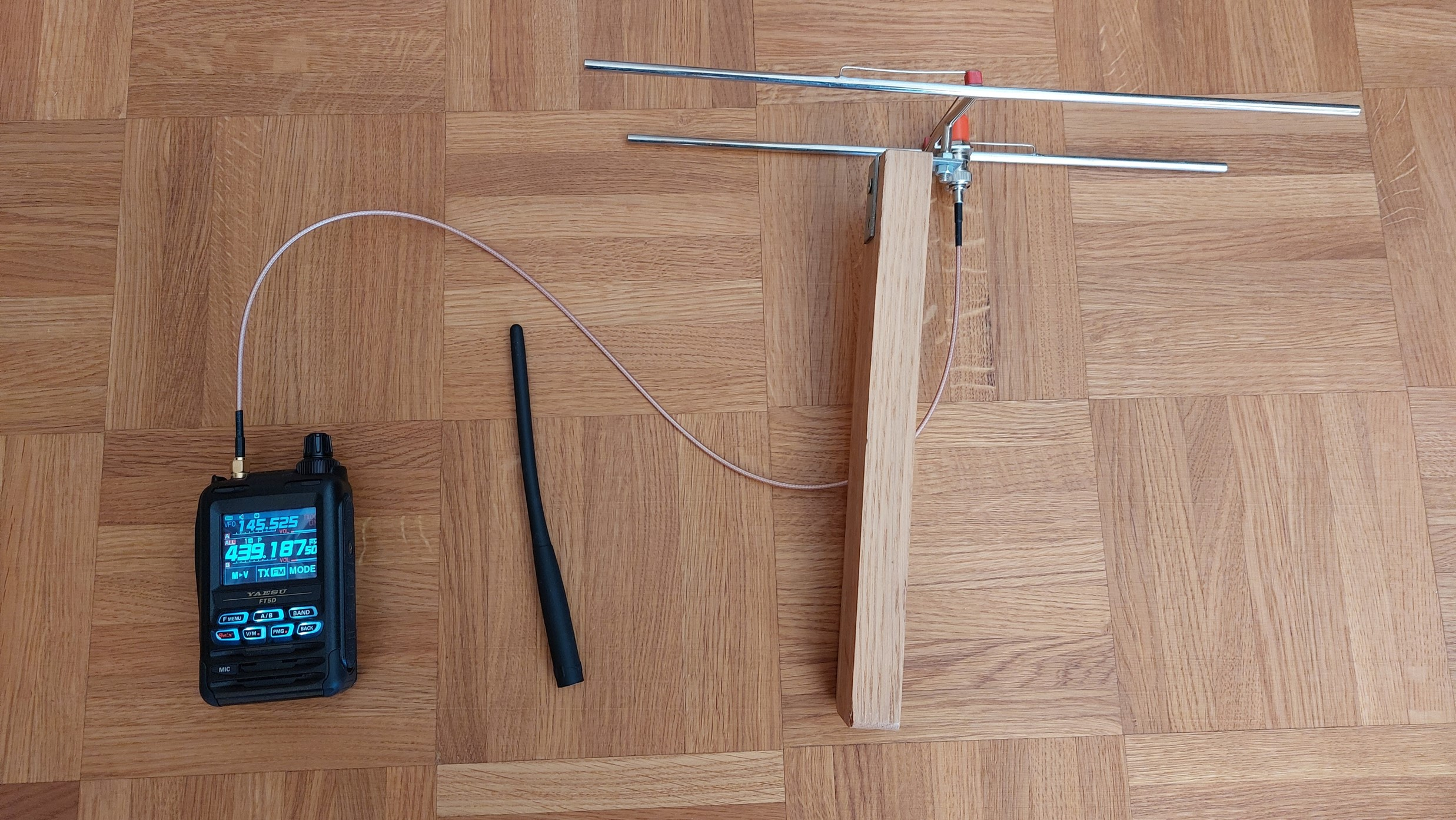 1) Kurzbeschreibung: Handfunkgerät, über ein dünnes Koaxialkabel mit einer kleinen, auf einem Holzstab montierten 2-Element-Richtantenne verbunden; neben dem Funkgerät eine Gummi-Stabantenne.

2) Ausführliche Beschreibung: Das Foto zeigt ein Handfunkgerät, das über ein dünnes Koaxialkabel mit einer kleinen, auf einem Holzstab montierten 2-Element-Richtantenne verbunden ist. Neben dem Funkgerät liegt eine abgenommene Gummi-Stabantenne.