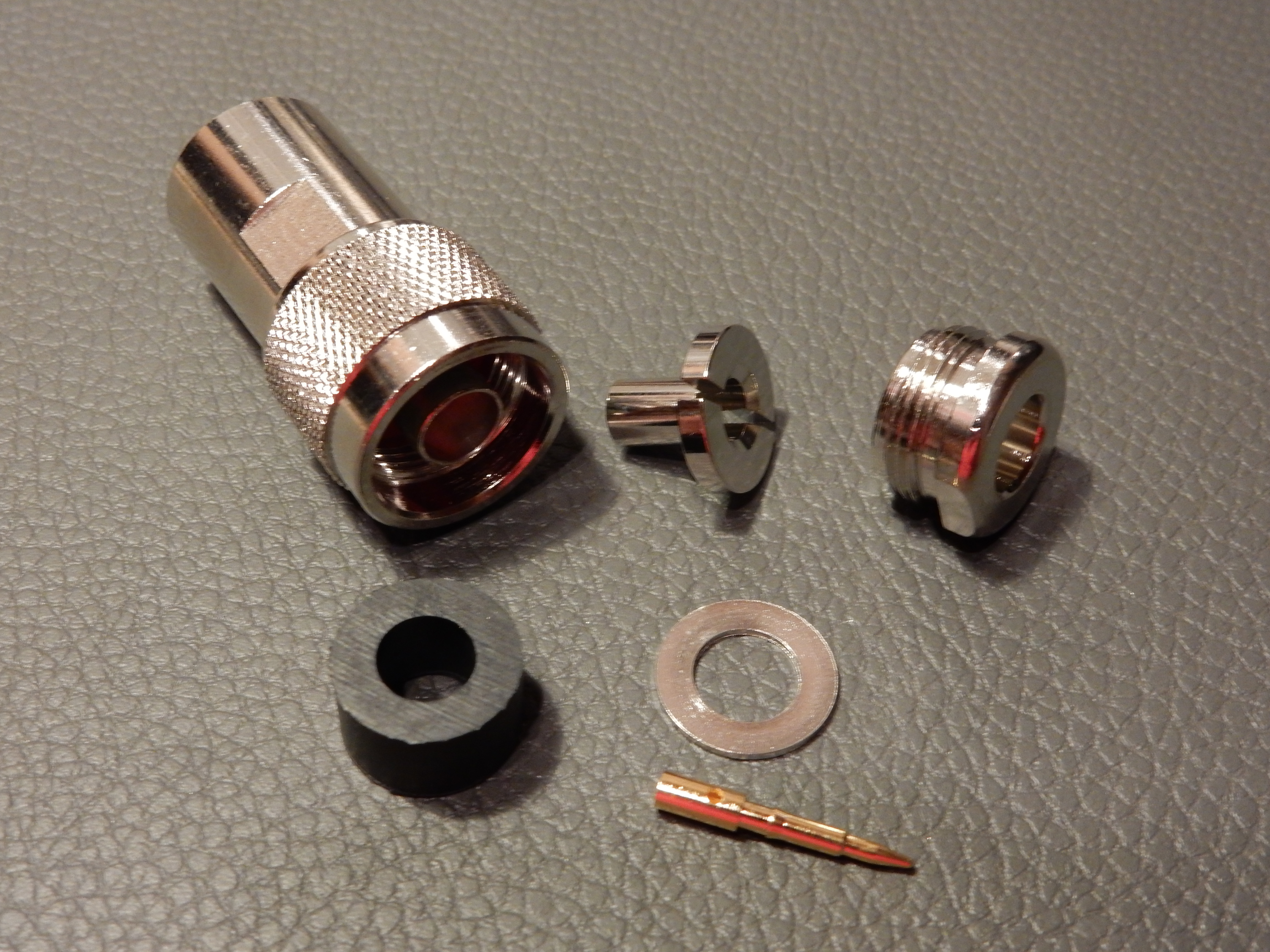 1) Kurzbeschreibung: In Einzelteile zerlegter N-Stecker.

2) Ausführliche Beschreibung: Das Foto zeigt einen in seine Einzelteile zerlegten N-Stecker. Links oben liegt das Steckergehäuse, rechts daneben die Kontakthülse sowie die Spannmutter mit Außengewinde. Links unten ist die Gummidichtung zu sehen, daneben der Druckring und darunter der Innenstift.