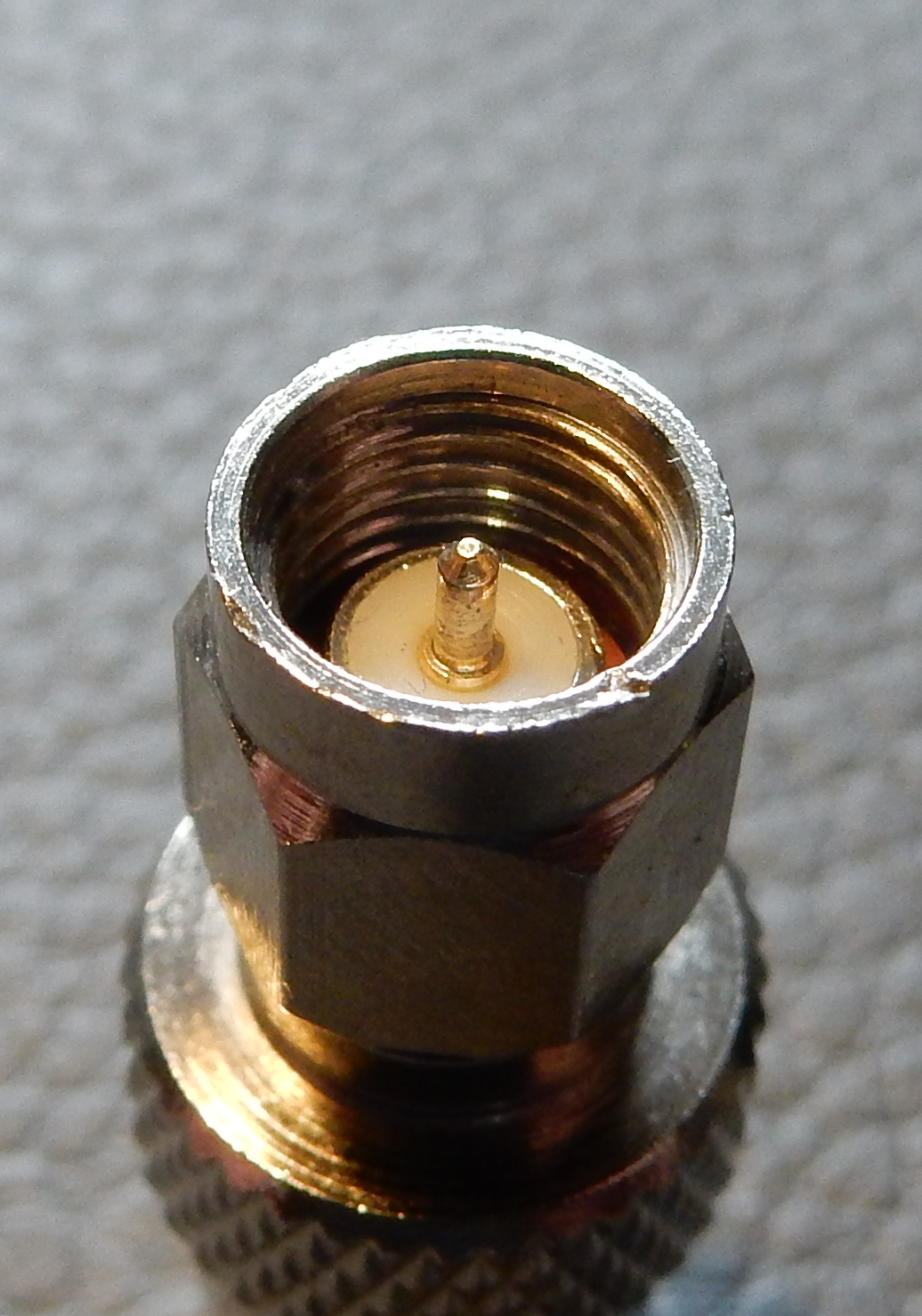 1) Kurzbeschreibung: Koaxialer SMA-Stecker mit vergoldetem Mittelkontakt, weißem Isolator und Innengewinde.

2) Ausführliche Beschreibung: Das Foto zeigt einen koaxialen SMA-Stecker mit vergoldetem Mittelkontakt, weißem Isolator und Innengewinde in einem Ring, an den sich nach unten ein sechseckiger Körper anschließt.