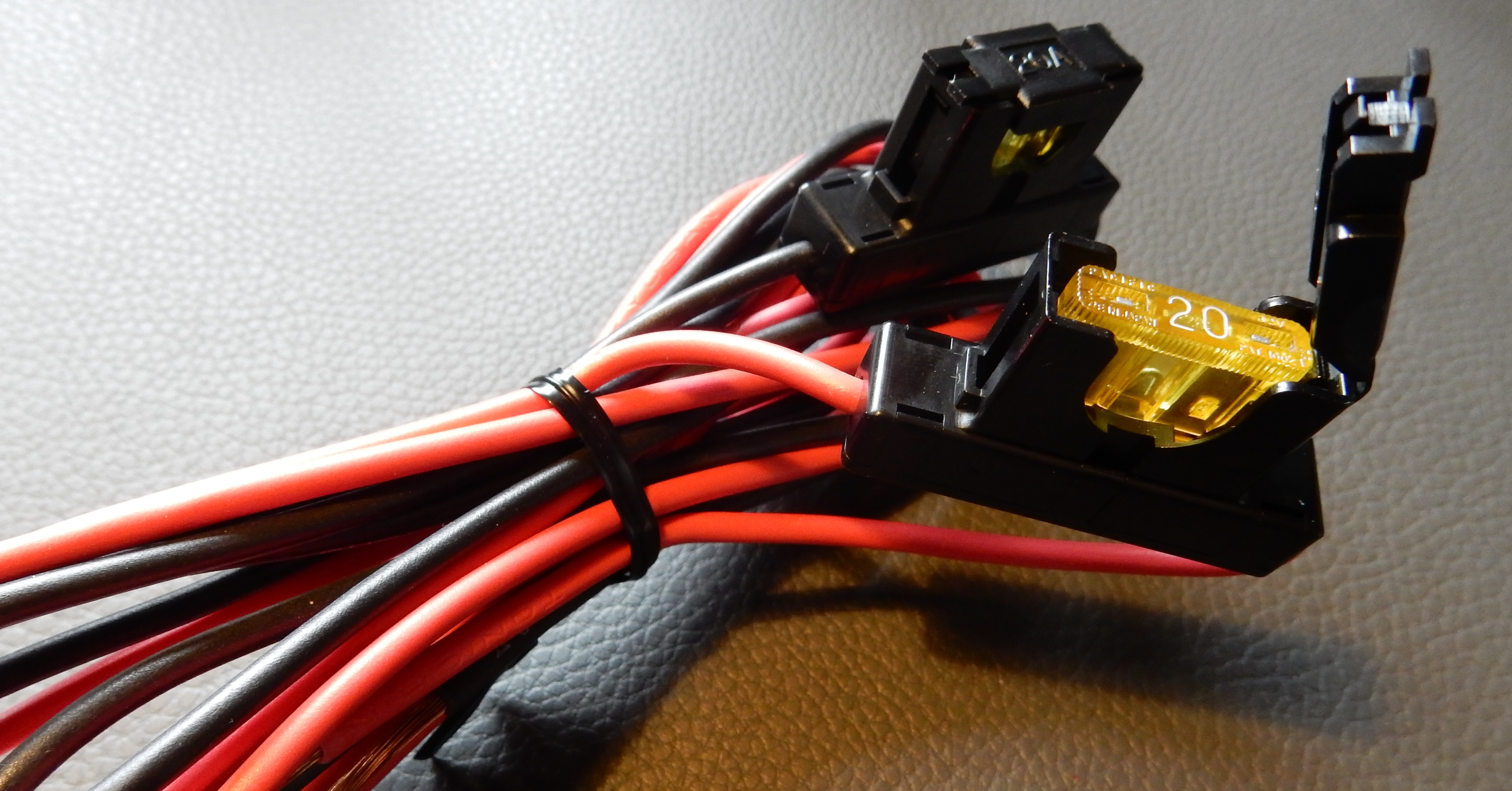 1) Kurzbeschreibung: Bündel roter und schwarzer Kabel mit zwei Inline-Sicherungen, von denen eine geöffnet ist und eine gelbe 20‑A‑Flachsicherung zeigt.

2) Ausführliche Beschreibung: Das Foto zeigt mehrere rote und schwarze Leitungen, die zu einem Bündel zusammengefasst und mit einem schwarzen Kabelbinder fixiert sind. Am rechten Bildrand sind zwei schwarze Sicherungshalter zu sehen. Einer ist aufgeklappt und gibt den Blick auf eine gelbe Flachstecksicherung mit der weißen Aufschrift „20“ frei, der andere Halter ist geschlossen.
