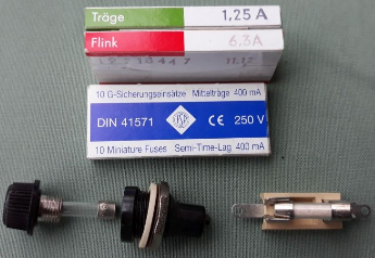1) Kurzbeschreibung: Foto mit drei Feinsicherungen mit unterschiedlicher Auslösezeit in Verpackungen sowie zwei Sicherungshalter.

2) Ausführliche Beschreibung: Das Foto zeigt drei Feinsicherungen mit unterschiedlicher Auslösezeit in Verpackungen, beschriftet mit „Träge“ (1,25 A), „Flink“ (6,3 A) und „Mittelträge“ (400 mA). Darunter sind zwei Sicherungshalter gezeigt. Links ist ein zerlegter Sicherungshalter mit Schraubgewinde zu sehen, in dem eine Feinsicherung aus Glas mit Metallkappen an den Enden steckt. Rechts davon befindet sich ein Sicherungshalter mit zwei Klemmen, die eine Feinsicherung aus Glas enthalten.