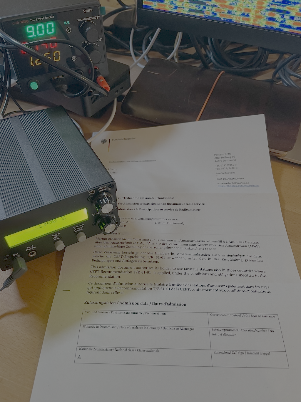 1) Kurzbeschreibung: Zulassungsbescheid vor dem Display eines Funkgerätes.

2) Ausführliche Beschreibung: Das Foto zeigt den Zulassungsbescheid zur Teilnahme am Amateurfunkdienst neben einem Funkgerät. Im Hintergrund ist ein Labornetzteil und ein Wasserfalldiagramm auf einem Computerbildschirm zu sehen.