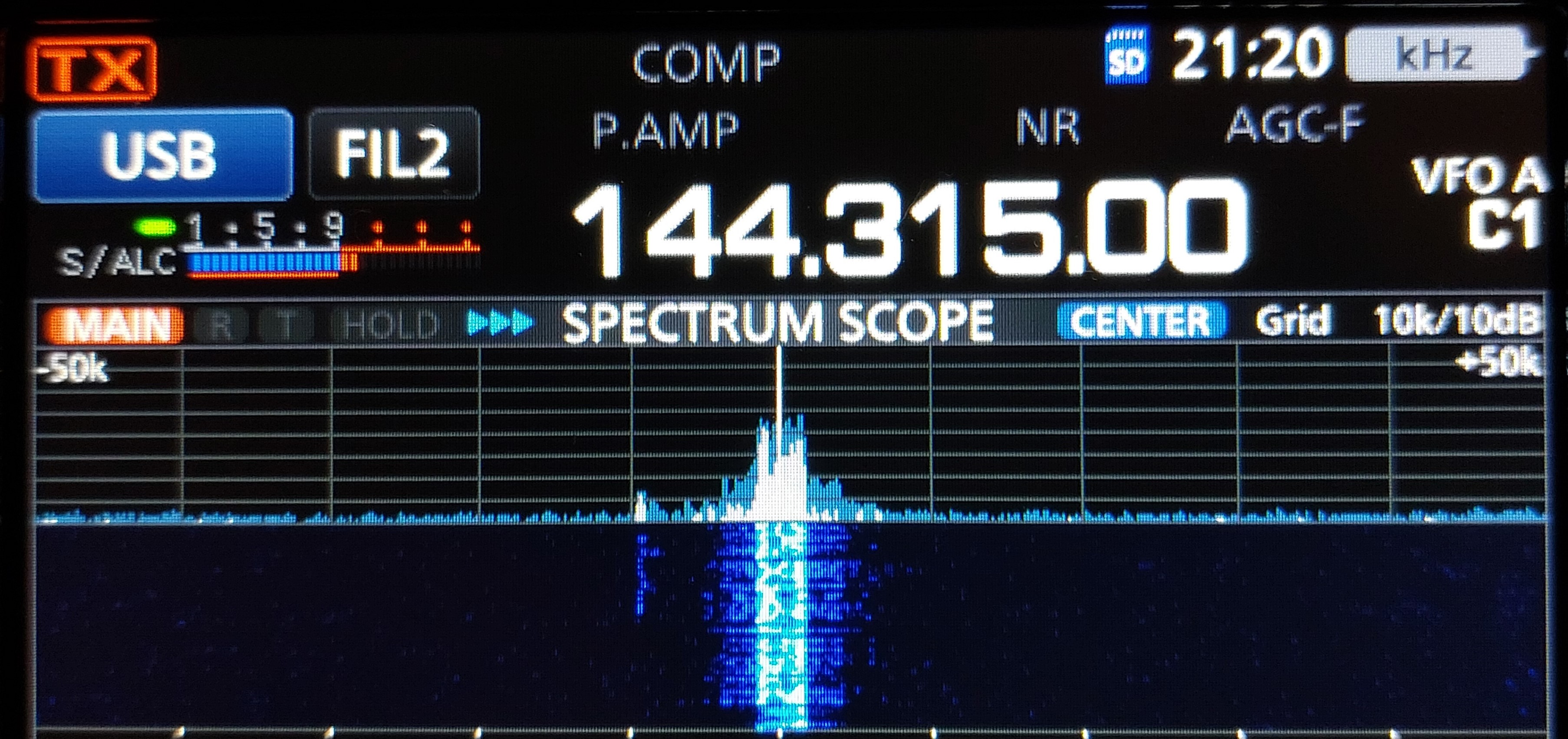 1) Kurzbeschreibung: Display eines Funkgerätes mit Frequenzanzeige „144.315.00“, Spektrums- und Wasserfalldarstellung eines starken Signals in der Mitte.

2) Ausführliche Beschreibung: Die Abbildung zeigt das Display eines Funkgerätes auf schwarzem Untergrund. Neben weiteren Beschriftungen steht in der Mitte in großen, weißen Ziffern die Frequenz „144.315.00“. Darunter ist in blauer Farbe die Anzeige des Spektrums um die eingestellte Frequenz zu sehen, die von den Angaben „–50k“ (links) und „+50k“ (rechts) begrenzt wird. In der Mitte gibt es einen schmalen, hohen Peak mit kleineren Flanken. Darunter befindet sich ein Wasserfalldiagramm auf dunkelblauem Hintergrund mit einer hellblau-weißen vertikalen Spur unterhalb des Peaks.