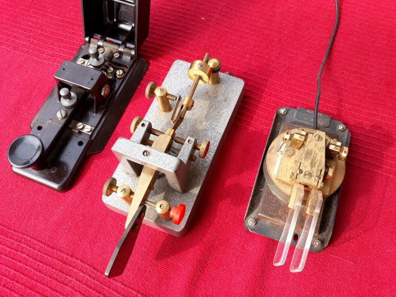 1) Kurzbeschreibung: Drei Morsetasten unterschiedlicher Bauform: Handtaste, halbautomatische Taste, Squeeze-Taste.

2) Ausführliche Beschreibung: Das Foto zeigt drei Morsetasten unterschiedlicher Bauform. Links steht eine Handtaste mit einem Tasthebel, Schrauben zur Hubeinstellung und zum Tastendruck und einem aufgeklappten Deckel. In der Mitte steht eine halbautomatische Taste mit einem senkrecht stehenden Tasthebel und einem Gewicht und einer Feder zur Rückstellung des Hebels nach dessen Betätigung sowie Stellschrauben. Rechts ist eine Squeeze-Taste mit zwei einander gegenüber stehenden Hebelarmen und Stellschrauben zu sehen.