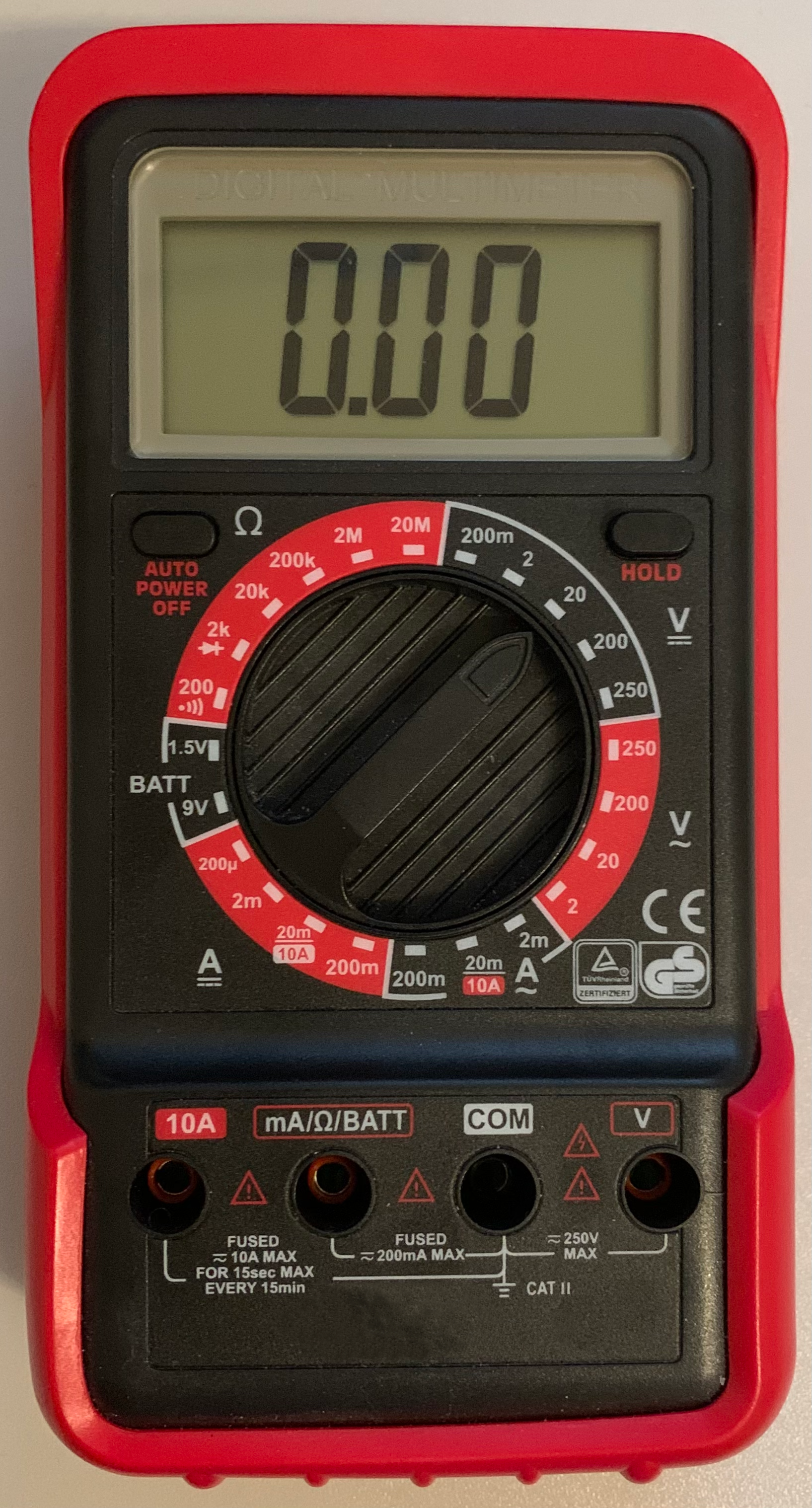 1) Kurzbeschreibung: Digitales Handmultimeter in roter Schutzhülle mit Anzeige „0.00“ und zentralem Drehwahlschalter.

2) Ausführliche Beschreibung: Das Handmultimeter besitzt oben ein graues LCD, das „0.00“ anzeigt. Darüber ist „DIGITAL MULTIMETER“ eingeprägt. Links unterhalb des Displays sitzt ein kleiner rechteckiger Taster mit der Beschriftung „AUTO POWER OFF“, rechts ein gleichartiger Taster „HOLD“. In der Mitte dominiert ein großer, geriffelter Drehwahlschalter, umgeben von einer Skala mit rotem Segment und weißen Markierungen. Es gibt die folgenden Messbereiche: Widerstand (Ω); Gleichspannung (V⎓); Wechselspannung (V~); Wechselstromstärke (A~); Gleichstromstärke (A⎓); Batteriespannung „BATT“. Rechts unterhalb des Drehwahlschalters sind die Konformitätszeichen „CE“, „TÜV Rheinland“ und „GS“ zu sehen. Unten gibt es vier Buchsen mit Beschriftungen von links nach rechts: „10A“, „mA/Ω/BATT“, „COM“ und „V“, dazwischen jeweils Warnsymbole und verschiedene Aufdrucke. Das Gehäuse ist schwarz mit einer roten, gummiartigen Umrandung.