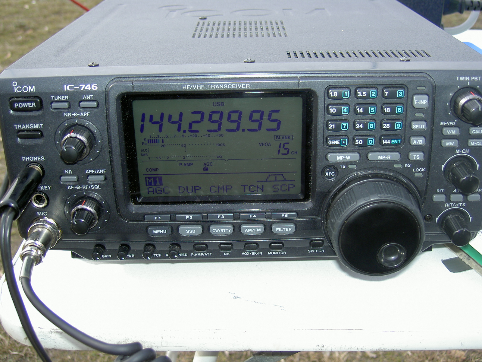 Der folgende Alt-Text wurde noch nicht geprüft: 1. Zusammenfassung: Frontansicht eines eingeschalteten Icom IC‑746 HF/VHF‑Transceivers auf einem weißen Tisch mit großer Frequenzanzeige 144.299.95.

2. Detaillierte Beschreibung: Das Gerät füllt das Bild, oben links steht das Logo „ICOM“ und die Modellbezeichnung „IC‑746“, darüber Lüftungsschlitze im Gehäusedeckel. Mittig befindet sich ein großes, beleuchtetes LCD; es zeigt links diverse Balkenanzeigen (u. a. S‑Meter/ALC/SWR), oben rechts den Betriebsmodus „USB“ und groß in Ziffern die Frequenz „144.299.95“. Im Display sind außerdem kleine Statusfelder wie „VFO A“ und „15 CH“ sowie Textzeilen mit Abkürzungen (z. B. „AGC“, „DUP“, „CMP“, „TCN“, „SCP“) erkennbar. Links vom Display sitzen Tasten und Drehregler mit Beschriftungen wie „POWER“, „TUNER“, „TRANSMIT“, „NR“, „APF/ANF“ und „AF‑RF/SQL“; darunter sind Frontbuchsen mit Aufschriften „PHONES“, „KEY“ und „MIC“, wobei am MIC‑Anschluss ein Kabel mit Metallstecker eingesteckt ist. Unterhalb des Displays verläuft eine Reihe Funktionstasten (u. a. „F1“ bis „F5“, „MENU“, „SSB“, „CW/RTTY“, „AM/FM“, „FILTER“) sowie kleinere Schalter und Regler. Rechts vom Display befindet sich ein Tastenfeld mit hellblau hinterlegten Band- und Funktionstasten, darunter Beschriftungen wie „1.8“, „3.5“, „7“, „14“, „21“, „28“, „50“, „144“, „SPLIT“, „A/B“, „V/M“. Rechts außen dominiert ein großer, geriffelter Abstimmknopf; daneben weitere Bedientasten und Drehregler wie „RIT/ΔTX“, „LOCK“, „MW“ und „M‑CH“. Das Gerät steht auf einem weißen Tisch; im unscharfen Hintergrund sind erdige/grasige Farbtöne zu erkennen.