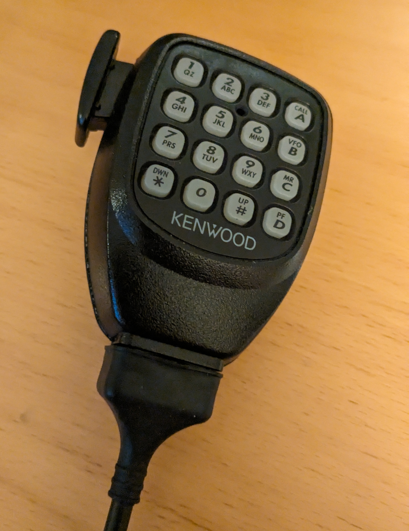 Der folgende Alt-Text wurde noch nicht geprüft: Kurzfassung: Ein schwarzes, kabelgebundenes Handgerät mit 4x4-Tastenfeld und Kenwood-Logo liegt auf einer hellen Holzoberfläche.

Detailbeschreibung: Das Gerät hat ein muschelförmiges, strukturiertes Kunststoffgehäuse in Schwarz. Auf der Vorderseite befindet sich ein gummiertes, hellgraues Tastenfeld in einem 4x4-Raster. Die ersten drei Spalten zeigen Zifferntasten 1–9 mit kleinen Buchstabenangaben darunter (1 QZ, 2 ABC, 3 DEF, 4 GHI, 5 JKL, 6 MNO, 7 PRS, 8 TUV, 9 WXY). In der rechten Spalte sind vier Tasten mit Aufdruck „CALL A“, „VFO B“, „MR C“ und unten „PF D“. In der untersten Reihe stehen links „*“ mit dem Zusatz „DWN“, in der Mitte „0“ und rechts „#“ mit dem Zusatz „UP“. Unterhalb des Tastenfeldes ist in weißen Großbuchstaben „KENWOOD“ aufgedruckt. Links am Gehäuse ragt ein kleiner, flacher Clip oder Haken hervor. Unten führt ein schwarzes Kabel mit Knickschutz aus dem Gerät. Der Hintergrund ist eine glatte, hellholzfarbene Tischplatte.