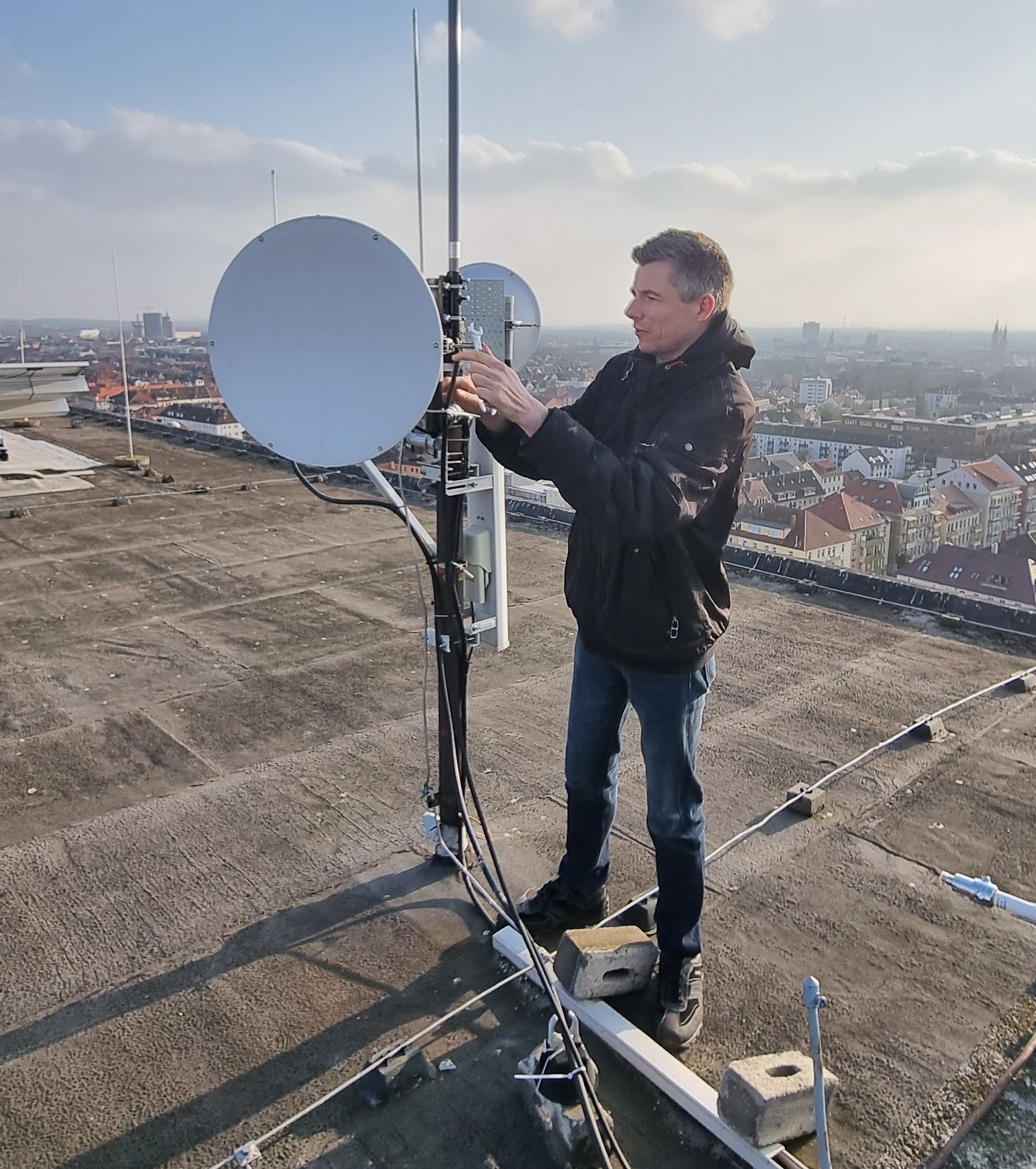 Der folgende Alt-Text wurde noch nicht geprüft: 1. Kurze Zusammenfassung: Eine Person arbeitet auf einem Flachdach an einer Funkanlage mit Parabolspiegel und Stabantennen, im Hintergrund eine weitläufige Stadtansicht unter leicht bewölktem Himmel.

2. Detaillierte Beschreibung: Im Vordergrund steht eine erwachsene Person in dunkler Jacke und Jeans auf einem bituminierten Flachdach neben einem Antennenmast. Am Mast sind ein großer, grauer runder Parabolspiegel, eine kleinere runde Antenne, eine gitterförmige Richtantenne sowie mindestens zwei vertikale Stabantennen montiert. Mehrere schwarze und weiße Koaxkabel verlaufen vom Mast nach unten und sind am Gestell fixiert; die Person hält die Hände an einer Halterung oder einem Anschluss am Mast. Der Mast ist an einer weißen Schiene befestigt, die mit grauen Betonsteinen beschwert ist; weitere Kabel und Verbinder liegen auf dem Dach. Die Sonne steht tief und wirft lange Schatten. Im Hintergrund sind Dächer und mehrstöckige Gebäude einer Stadt, einzelne Hochhäuser und Kirchtürme sowie ein weiter, diesiger Horizont unter locker bewölktem Himmel zu sehen.