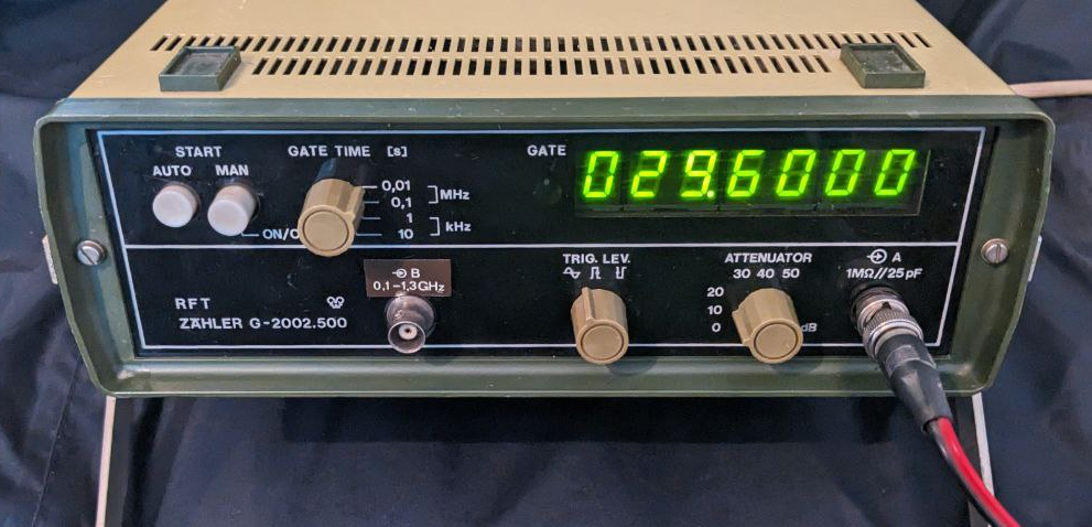 1) Kurzbeschreibung: Frontansicht eines Frequenzzählers mit leuchtend grünem 7‑Segment‑Display und der Anzeige „029.6000“, mehreren Drehknöpfen, Tastern und BNC‑Anschlüssen.	

2) Ausführliche Beschreibung: Das Foto zeigt die Frontansicht eines Frequenzzählers mit Metallgehäuse mit Lüftungsschlitzen oben. Die Frontplatte ist schwarz mit weißer Beschriftung und rundum von einem grünen Rahmen eingefasst. Es gibt mehrere Drucktasten und Drehknöpfe. Oben rechts gibt es ein grünes 7-Segment-Display mit der Anzeige „029.6000“.