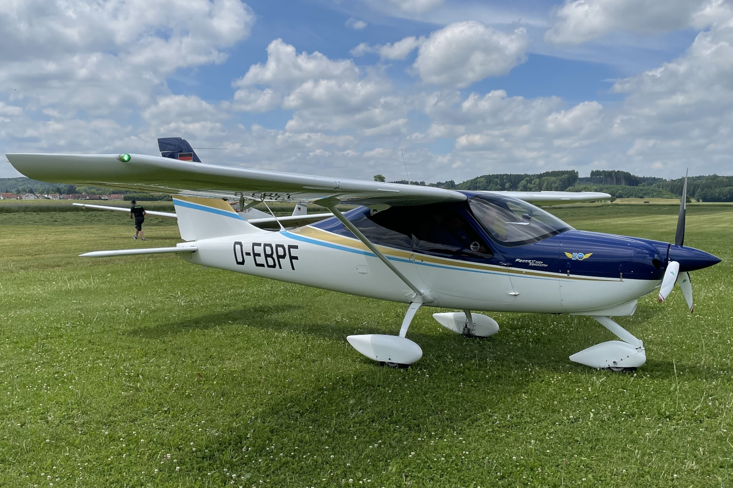 Der folgende Alt-Text wurde noch nicht geprüft: 1. Kurze Zusammenfassung: Ein kleines einmotoriges Hochdecker-Flugzeug steht auf einer grasigen Flugplatzfläche unter einem leicht bewölkten Himmel.

2. Detaillierte Beschreibung: Das Flugzeug ist überwiegend weiß, die Front mit Motorhaube und Kabinenbereich ist dunkelblau; entlang des Rumpfs verlaufen goldene und hellblaue Zierstreifen. Auf der linken Rumpfseite steht groß das Kennzeichen „D-EBPF“. Am Bug sitzt ein stillstehender zweiblättriger Propeller mit weißem Spinner; an einem Propellerblatt hängt ein rotes Band. Die Maschine hat ein Bugradfahrwerk mit Verkleidungen an allen drei Rädern. Die Tragfläche ist oben am Rumpf montiert und mit Streben abgestützt; an der linken Flügelspitze leuchtet ein grünes Positionslicht. Durch die getönte Kabinenhaube ist eine Person im Cockpit zu erkennen. Auf der Motorhaube ist ein kleines „50“-Abzeichen zu sehen, am Seitenleitwerk eine kleine Flagge. Im Hintergrund befinden sich eine Person zu Fuß, eine weitläufige Wiese, Bäume und sanfte Hügel; darüber ziehen zahlreiche weiße Wolken.