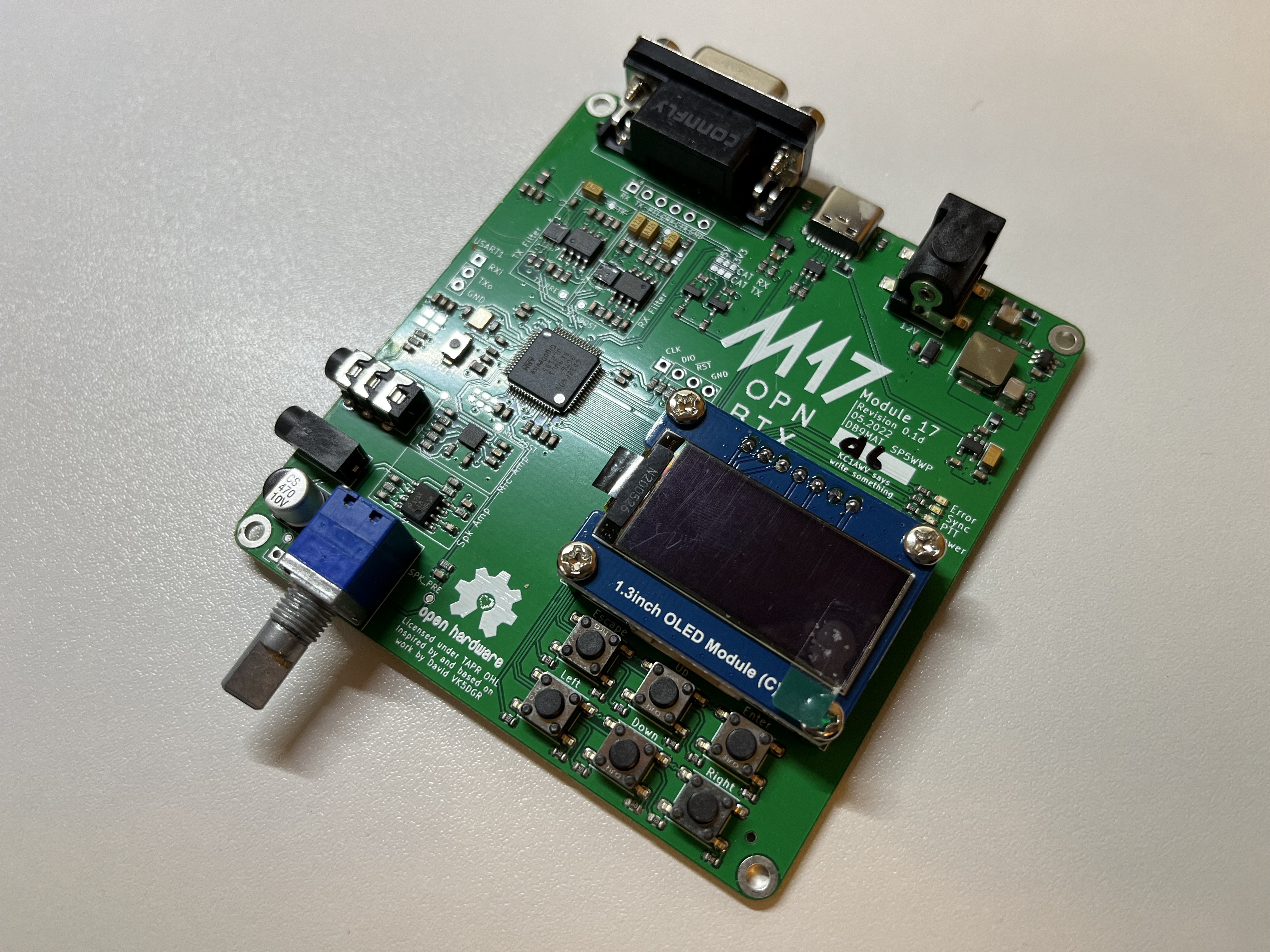Der folgende Alt-Text wurde noch nicht geprüft: 1) Kurze Zusammenfassung: Eine grüne Elektronikplatine mit 1,3‑Zoll‑OLED‑Display, mehreren Tastern, einem Drehgeber und diversen Anschlüssen, schräg auf einer hellen Oberfläche aufgenommen.

2) Detaillierte Beschreibung: Die rechteckige Leiterplatte zeigt oben groß den Aufdruck „M17“ sowie weitere Beschriftungen wie „OPN“ und „Module 1.7“. Im oberen Bereich sitzt mittig eine 9‑polige D‑Sub‑Buchse; rechts oben befindet sich eine schwarze Hohlstecker‑Strombuchse, daneben mehrere SMD‑Bauteile. In der Mitte ist ein aufgeschraubtes, blau gerahmtes Modul mit der Beschriftung „1.3 inch OLED Module (C)“ zu sehen, das über einen Stiftleisten‑Header verbunden ist. Unterhalb des Displays liegen fünf runde Taster in zwei Reihen, beschriftet mit „Left“, „Right“, „Up“, „Down“ und „Select“. Links unten ragt der Metallschaft eines blauen Drehgebers (Encoder) aus der Platine. Über die Fläche verteilt befinden sich integrierte Schaltkreise, Widerstände, Kondensatoren, ein kleiner Schiebeschalter, unbestückte Löt‑ und Stiftleistenplätze sowie vier Befestigungslöcher an den Ecken. Die Platine liegt frei auf einer leicht strukturierten, hellen Oberfläche, die weiche Schatten wirft.
