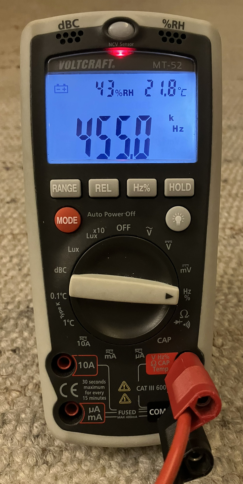 Dieser Alt-Text wurde noch nicht überprüft.

Kurzfassung: Ein tragbares Digitalmultimeter mit blau beleuchtetem Display zeigt 455.0 kHz sowie 43 % relative Luftfeuchte und 21.8 °C an.

Detaillierte Beschreibung: Das Gerät im grauschwarzen Gummischutzgehäuse liegt auf einem beigen Teppich. Oben leuchtet eine rote LED neben der Aufschrift „NCV Sensor“; darüber sind die Hinweise „dBC“ und „%RH“ mit kleinen Lüftungsöffnungen zu sehen. Auf dem Display steht links ein Batteriesymbol, daneben „43%RH“ und rechts „21.8°C“; die großen Ziffern in der Mitte zeigen „455.0“, flankiert von den Einheiten „k“ und „Hz“. Unter dem Display befinden sich Tasten mit den Beschriftungen „RANGE“, „REL“, „Hz%“ und „HOLD“, links eine rote „MODE“-Taste und rechts eine Taste mit Lampensymbol. Der zentrale Drehschalter steht auf der rechten Seite in der Position mit der Beschriftung „Hz %“; weitere Skalen rundherum sind u. a. mit „V“, „mV“, „CAP“ und „Lux“ markiert. Unten sind vier Buchsen zu sehen: „10A“, „µA mA“, „COM“ und „V Hz Ω CAP Temp“; in „COM“ steckt ein schwarzer Stecker, in der rechten Buchse ein roter Stecker mit Kabel, daneben Sicherheits- und Messkategorienhinweise wie „CAT III 600V“ und „FUSED MAX 400mA“. Auf der Front sind außerdem die Markierungen „VOLTCRAFT“ und „MT-52“ sowie „Auto Power Off“ sichtbar.