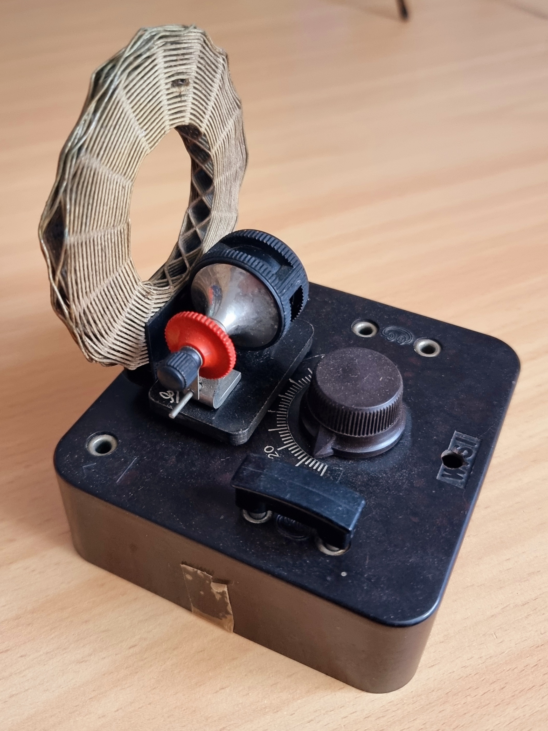 Der folgende Alt-Text wurde noch nicht geprüft: Kurzfassung: Ein schwarzes, älter wirkendes Tischgerät mit senkrechter Ringspule und mehreren Einstellelementen steht auf einer hellen Holzoberfläche.

Detaillierte Beschreibung: Auf einem rechteckigen, dunkelbraunen/ schwarzen Gehäuse (mit abgenutzter Oberfläche) sitzt links eine flache, aufrecht stehende Ringspule aus eng gewickeltem, hellem Drahtband mit rautenförmigem Muster. Davor befinden sich ein kleiner schwarzer Einstellmechanismus mit rotem gerändelten Rädchen und ein glänzendes, halbkugeliges Metallteil. Rechts ist ein großer, gerändelter Drehknopf neben einer halbkreisförmigen Skala mit Markierungen (u. a. 0–20), darüber ein schwarzer Schieber als Zeiger. In der rechten oberen Gerätefläche liegen zwei versenkte Metallschrauben/Anschlüsse; an zwei Ecken sind Bohrungen sichtbar. An der Frontseite klebt ein kleines Stück vergilbtes Klebeband. Der Hintergrund ist unscharf, die Holzplatte zeigt feine Maserung.