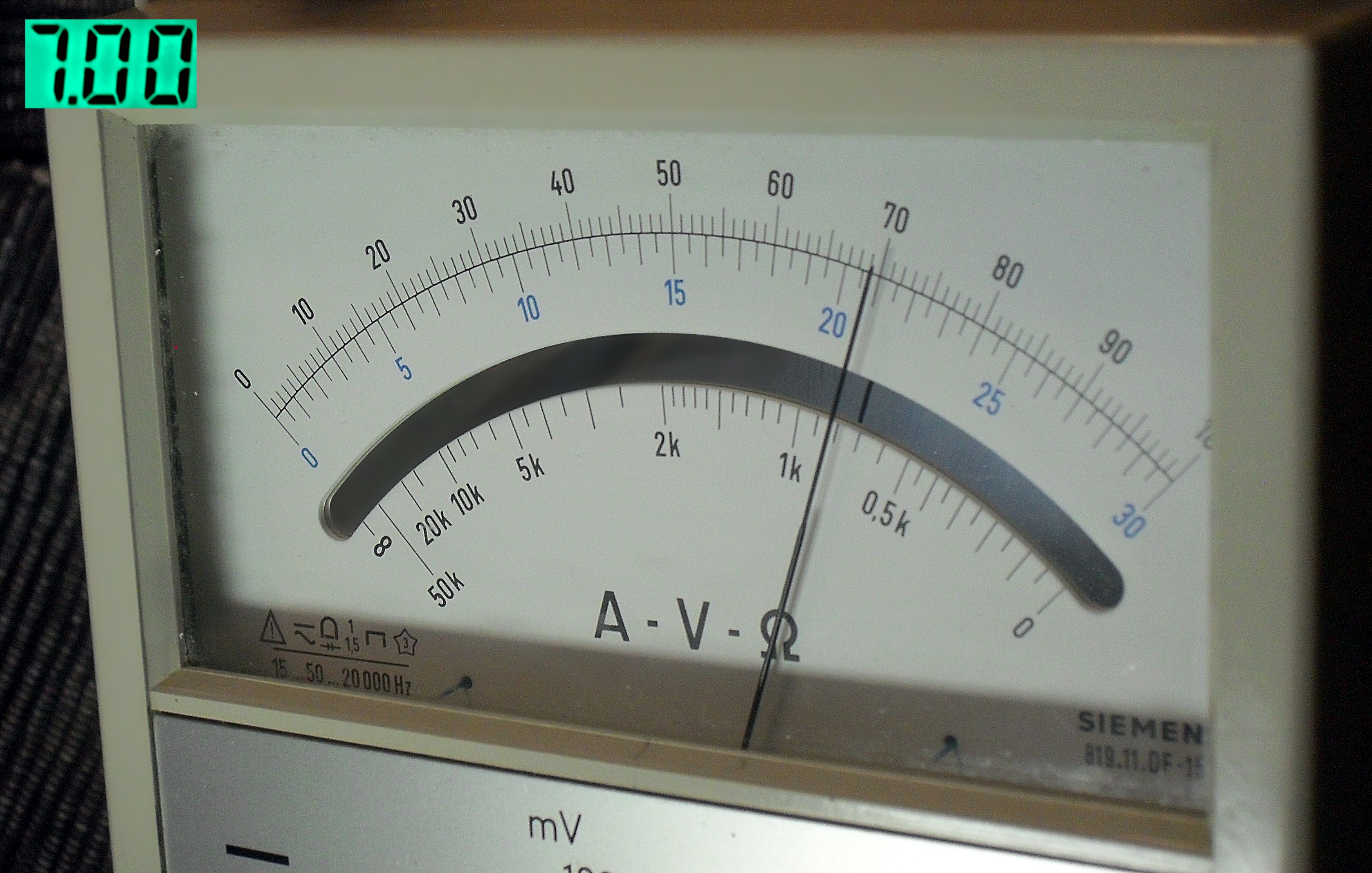 Der folgende Alt-Text wurde noch nicht geprüft: 1. Kurzzusammenfassung: Frontansicht einer analogen Messskala in einem beigen Gehäuse mit Zeigeranzeige und mehreren Skalen; der Zeiger steht nahe der 70 auf der äußeren Skala.

2. Detaillierte Beschreibung: Das Bild zeigt das Fenster eines analogen Messgeräts mit weißer Skalenfläche und halbkreisförmiger Mehrfachskala. Außen ist eine 0–90-Teilung mit feinen Strichen zu sehen, Zwischenwerte (z. B. 5, 10, 15, 20, 25, 30) sind in Blau markiert. Darunter verlaufen weitere Skalenbögen, u. a. eine mit logarithmischer Beschriftung in kΩ (0,5k, 1k, 2k, 5k, 10k, 20k, 50k). In der Mitte steht „A • V • Ω“. Ein schmaler Zeiger verläuft schräg nach oben rechts und zeigt knapp rechts von der 70 an. Unten links befinden sich kleine Piktogramme (u. a. ein Warnsymbol und eine „1,5“-Angabe) sowie der Text „15 • 50 • 20000 Hz“. Unten rechts ist ein teilweise sichtbarer „SIEMENS“-Schriftzug mit kleiner Modellbezeichnung. Das Gehäuse ist beige, der Skalenbereich durch Glas abgedeckt; am oberen linken Bildrand leuchtet ein grünes 7‑Segment‑Display mit „7.00“. Im linken Hintergrund ist ein dunkler, textiler oder gerippter Untergrund zu erkennen.