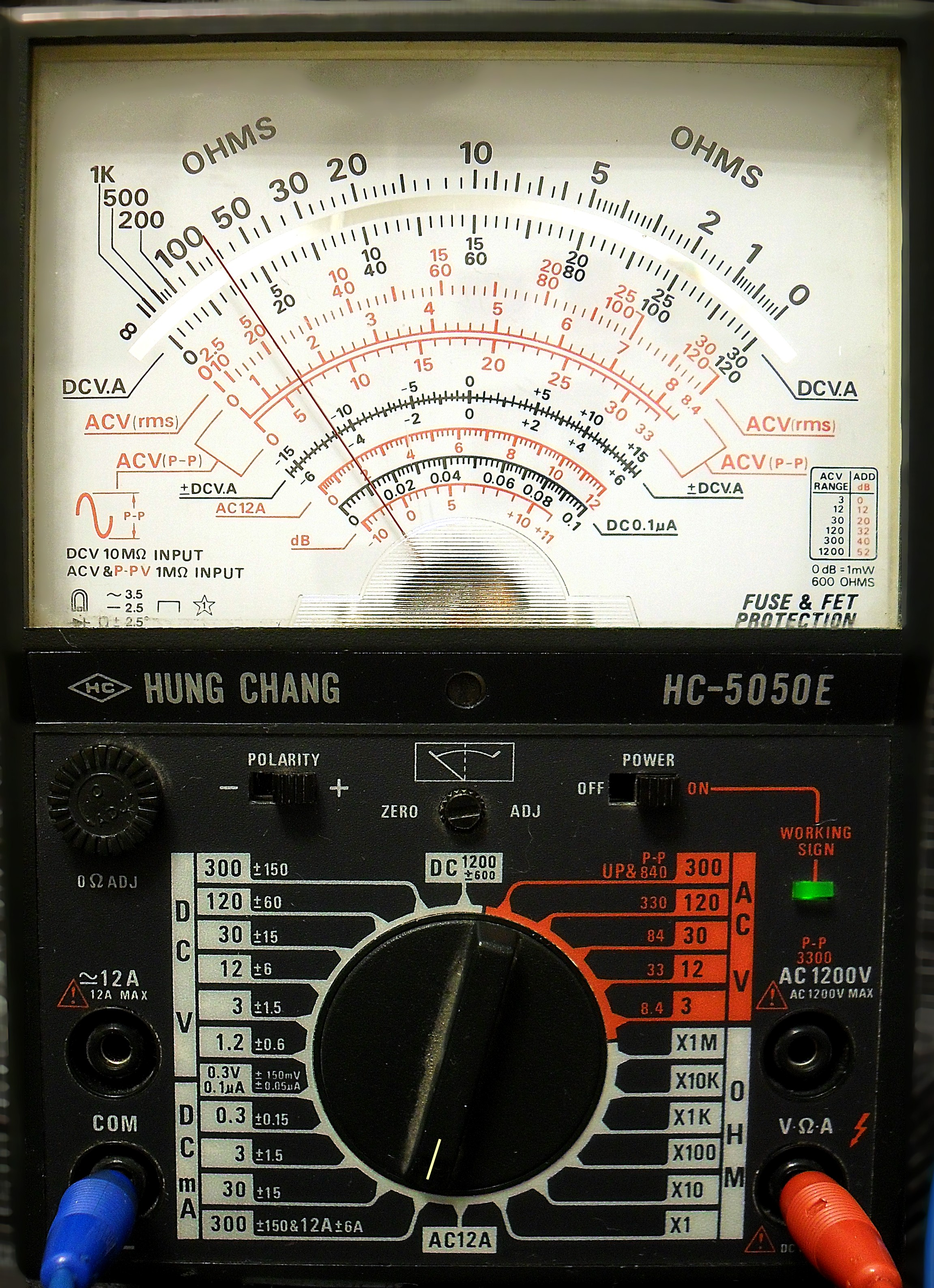 Dieser Alt-Text wurde noch nicht überprüft.

1. Zusammenfassung: Nahaufnahme eines analogen Multimeters mit großer Zeigerskala, zentralem Drehschalter und eingesteckten Messleitungen.

2. Detaillierte Beschreibung: Das Bild zeigt die Front eines Messgeräts „HUNG CHANG HC-5050E“. Oben dominiert eine große, halbkreisförmige Skala mit mehreren übereinanderliegenden Teilungen; außen sind „OHMS“-Markierungen, darunter rote und schwarze Skalen für DCV.A, ACV (rms), ACV (P‑P), dB und weitere Bereiche zu sehen. Ein Spiegelstreifen unter der Skala dient zur Parallaxekorrektur; der Zeiger ist sichtbar. Darunter befinden sich Bedienfelder mit Aufdrucken und Schaltern: links ein geriffelter „0Ω ADJ“-Knopf und ein kleiner Schiebeschalter „POLARITY“, mittig ein „ZERO/ADJ“-Regler, rechts ein Kippschalter „POWER“ auf „ON“ sowie eine leuchtende grüne Kontroll-LED mit der Beschriftung „WORKING SIGN“. Im unteren Bereich sitzt ein großer, schwarzer Drehwahlschalter, umgeben von weißen und orangenen Bereichsangaben für Gleich- und Wechselspannung, Gleichstrom und Widerstand (z. B. 300, 120, 30, 12, 3 sowie X1, X10, X100, X1K, X10K, X100K). Links unten sind zwei Buchsen mit Beschriftung „12A“ und „COM“, rechts unten eine Buchse „V·Ω·A“; in „COM“ steckt ein blaues Kabel, in „V·Ω·A“ ein rotes Kabel. Zahlreiche Skalen- und Bereichsangaben sind fein bedruckt; das Gehäuse ist dunkel und zeigt leichte Gebrauchsspuren.