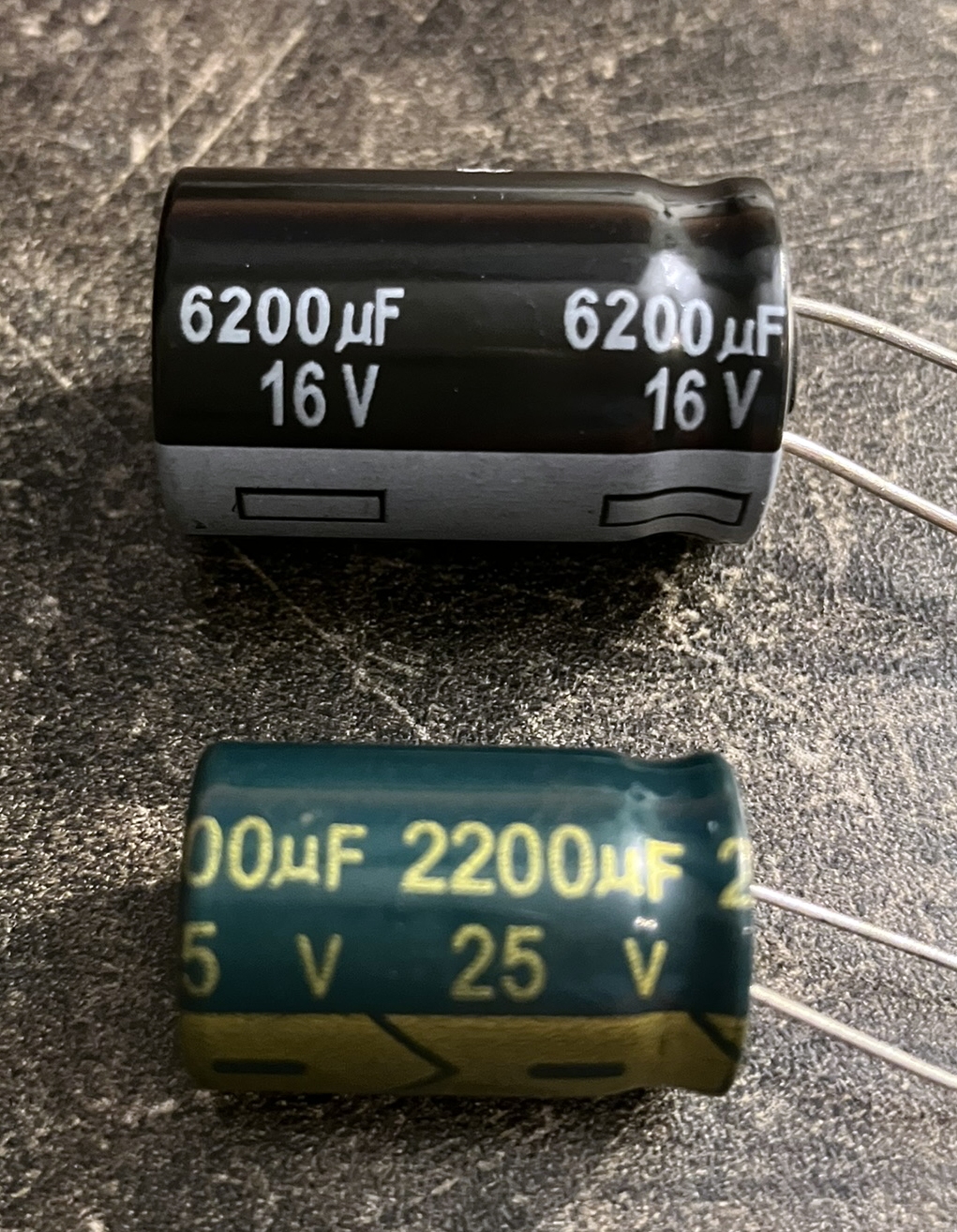 Der folgende Alt-Text wurde noch nicht geprüft: Kurzfassung: Zwei liegende Elektrolytkondensatoren mit Drahtanschlüssen auf einer dunklen, rauen Arbeitsfläche.

Detailbeschreibung: Oben ein größerer, zylindrischer Kondensator mit schwarzem Gehäuse und grauem Ring am unteren Ende; in weißer Schrift steht mehrfach „6200 µF 16 V“, die beiden Anschlussdrähte ragen nach rechts heraus. Darunter ein etwas kleinerer, dunkelgrüner Kondensator mit gelb-goldener Aufschrift „2200 µF 25 V“ und zusätzlichen Markierungen auf einem olivfarbenen Streifen; auch hier führen zwei Drähte nach rechts. Die Oberfläche zeigt Kratzer und Staub, das Licht erzeugt glänzende Reflexe auf den Kondensatoren, die nahezu parallel zueinander liegen.