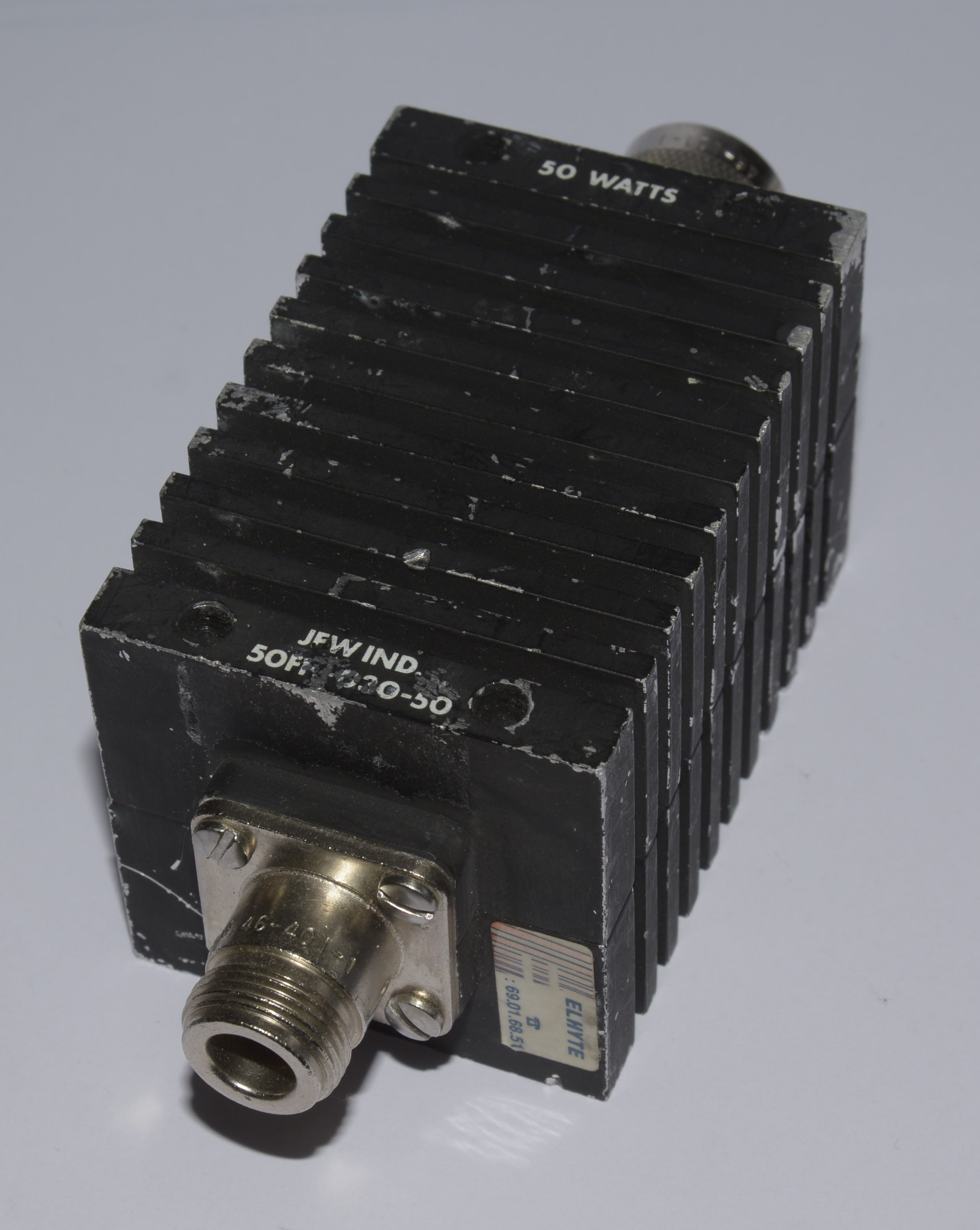Dieser Alt-Text wurde noch nicht überprüft.

Kurzfassung: Schwarzer, quaderförmiger Metallblock mit dichten Kühlrippen, der Aufschrift „50 WATTS“ und Koaxialanschlüssen an beiden Enden.

Detaillierte Beschreibung: Das Objekt liegt schräg auf einem hellgrauen Hintergrund und besteht aus einem schwarzen, gerippten Kühlkörper mit mehreren parallelen Längsrippen. An der Vorderseite sitzt ein koaxialer Einbausteckverbinder auf einer rechteckigen Metallflanschplatte, befestigt mit zwei Schrauben; die Lackierung ist an Kanten und Flächen sichtbar angeschlagen. Auf der Stirnseite sind die Worte „JFW IND.“ sowie eine teils abgeriebene Modellbezeichnung zu lesen. Auf der Oberseite steht „50 WATTS“. Auf der rechten Seitenfläche klebt ein kleiner Aufkleber mit der Aufschrift „ELMITE“ und einer Zahlenfolge. Hinten ist ein weiterer koaxialer Anschluss teilweise sichtbar. Das Licht fällt von oben links ein und erzeugt Schatten in den Rippenzwischenräumen.