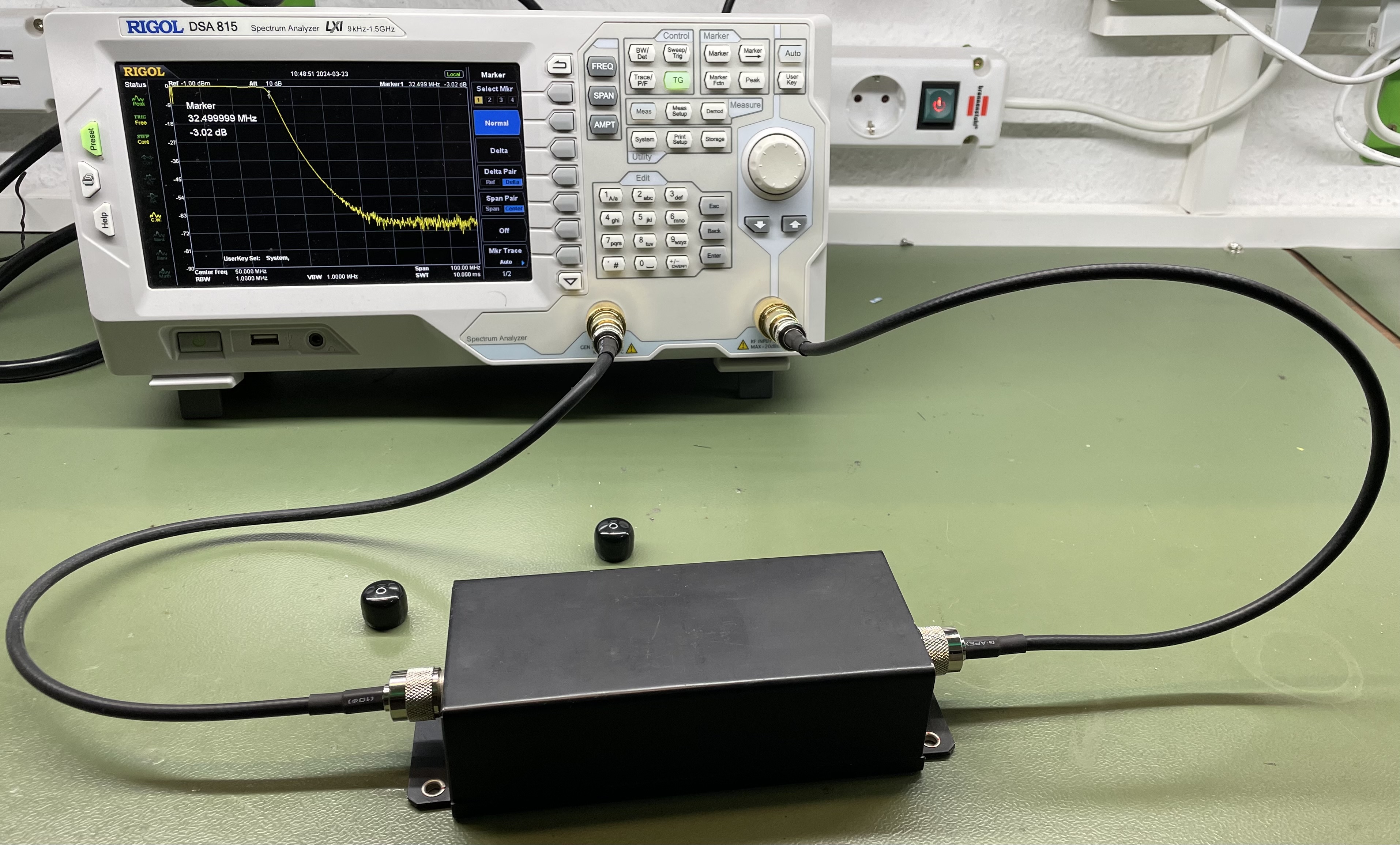 Der folgende Alt-Text wurde noch nicht geprüft: - Zusammenfassung: Ein Spektrumanalysator misst ein schwarzes, rechteckiges HF-Gehäuse auf einer Laborwerkbank über zwei Koaxkabel.

- Detaillierte Beschreibung: Links steht ein weiß-grauer Rigol DSA815 (Beschriftung „Spectrum Analyzer 9 kHz–1.5 GHz“) mit Farbdisplay und vielen Tasten sowie einem großen Drehknopf. Auf dem Display ist ein schwarzes Messraster mit einer gelben Kurve zu sehen; ein Marker ist bei etwa 32,5 MHz eingeblendet und zeigt rund −3 dB an. Unten rechts am Gerät sind zwei koaxiale Anschlüsse belegt; von dort führen zwei schwarze Koaxkabel zu einem mattschwarzen, rechteckigen Metallgehäuse mit Koaxbuchsen an beiden Stirnseiten und Befestigungslaschen, das vorne auf der grünlichen Tischoberfläche liegt. Auf dem Tisch liegen außerdem zwei kleine, runde schwarze Kappen. Im Hintergrund sind eine Steckdose, ein Kippschalter mit Leuchte und Kabelkanäle an der Wand zu sehen.