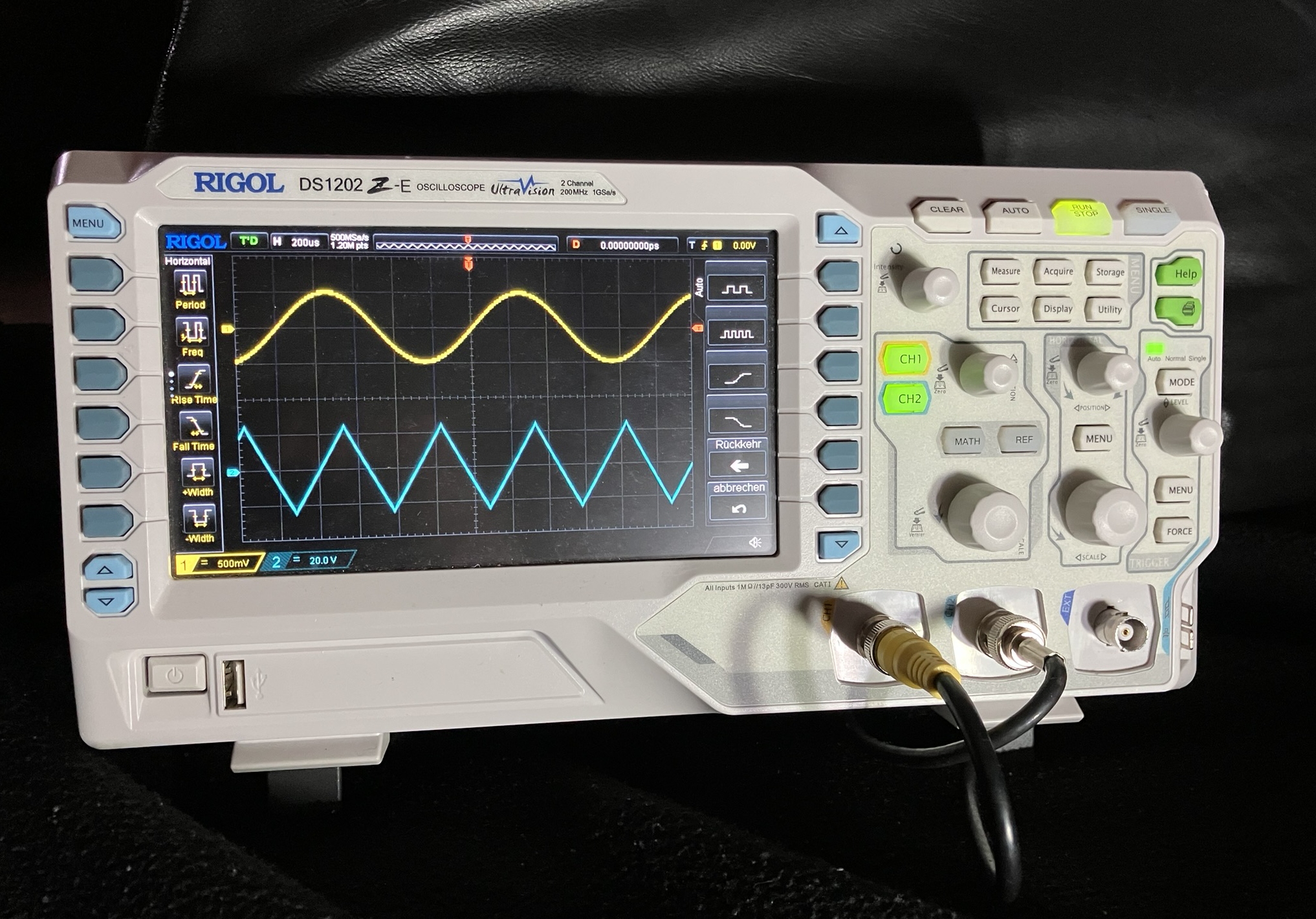 Der folgende Alt-Text wurde noch nicht geprüft: 1) Zusammenfassung: Ein digitales Rigol-Oszilloskop zeigt gleichzeitig eine gelbe Sinuskurve und eine blaue Dreieckskurve auf seinem Display.

2) Detaillierte Beschreibung: Das Gerät ist ein weißes Tischoszilloskop mit der Aufschrift „RIGOL DS1202Z‑E“ und „UltraVision“, fotografiert auf dunklem Hintergrund. Auf dem farbigen Bildschirm ist ein schwarzgraues Raster mit zwei Messkurven zu sehen: oben eine gelbe, weich geschwungene Sinuslinie, darunter eine blaue, gleichmäßig gezackte Dreieckslinie; links und rechts vom Messfeld stehen Symbolleisten mit gelben Funktionspiktogrammen (z. B. Periodendauer, Frequenz) und rechts vertikale Softkeys mit Kurven-Symbolen sowie deutschen Beschriftungen wie „Rückkehr“ und „abbrechen“. Am unteren Displayrand sind farbige Kanalbalken und Skalierungsangaben erkennbar. Rechts neben dem Display befinden sich zahlreiche Tasten und Drehregler; die Kanal-Tasten „CH1“ und „CH2“ leuchten grün. Unten rechts sind zwei BNC‑Buchsen mit einem gelben und einem schwarzen Koaxkabel belegt, eine weitere Buchse daneben ist frei. Links unten am Gerät sind ein Netzschalter und ein USB‑Anschluss sichtbar.