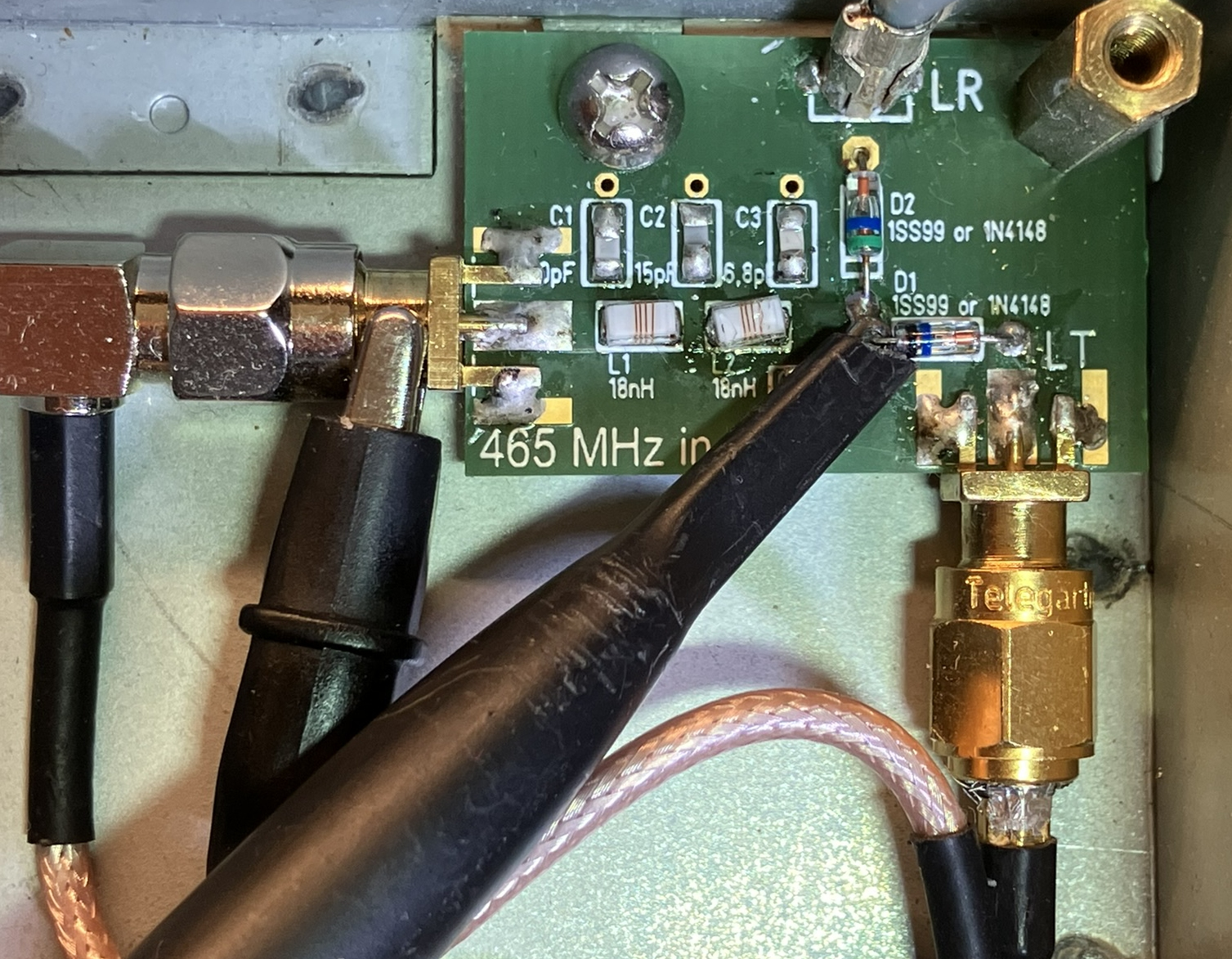 Dieser Alt-Text wurde noch nicht überprüft.

1) Kurzfassung: Geöffnete Elektronikbaugruppe in einem Metallgehäuse mit einer kleinen grünen Leiterplatte, mehreren Koax‑Steckverbindern und angesetzten Messleitungen.

2) Detaillierte Beschreibung: In der rechten Bildhälfte liegt eine grüne Leiterplatte mit weißer Beschriftung; sichtbar sind u. a. Bezeichnungen C1, C2, C3, L1, L2, D1, D2 sowie der Aufdruck „465 MHz in“. Neben den Markierungen für D1/D2 sind zwei kleine bedrahtete Bauteile mit Farbringen eingelötet; daneben liegen weitere bestückte und unbestückte SMD‑Plätze. Am rechten Rand ragt ein goldfarbener, verschraubter Koax‑Steckverbinder (SMA‑Bauform) nach unten aus dem Gehäuse; auf dem Sechskant steht „Telegärtner“. Links führt ein schwarzes Koaxkabel über einen rechtwinkligen metallischen Adapter auf die Platine. Über der Platine sind zwei schwarze Tastköpfe oder Leitungen angesetzt; ein spitzer schwarzer Prüfstift berührt einen Lötpunkt neben den beschrifteten Bauteilen. Die Platine ist mit mehreren Lötstellen, etwas Flussmittelrückständen und einer Schraube am Gehäuse befestigt; der Hintergrund besteht aus blankem Metall. Ein rosafarben ummanteltes Koaxkabel verläuft von unten rechts zum goldfarbenen Stecker.