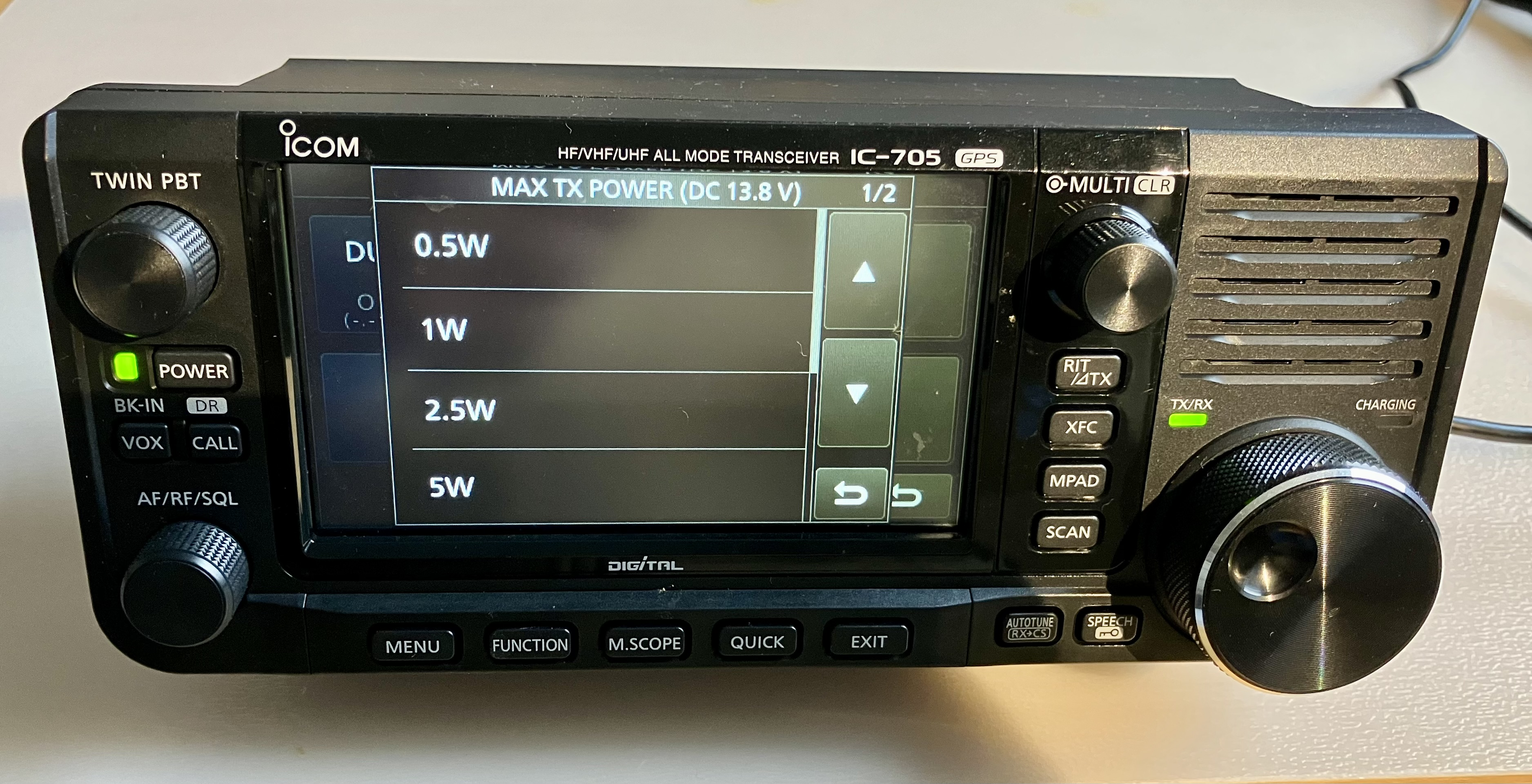 Dieser Alt-Text wurde noch nicht überprüft.

1) Kurze Zusammenfassung: Frontansicht eines ICOM IC-705 Transceivers mit eingeschaltetem Farbdisplay, das das Menü „MAX TX POWER (DC 13.8 V)“ mit wählbaren Sendeleistungen zeigt.

2) Detaillierte Beschreibung: Das Gerät steht auf einer hellen Oberfläche; oben auf der Frontblende sind die Aufdrucke „ICOM“, „HF/VHF/UHF ALL MODE TRANSCEIVER IC-705“ und „GPS“ sichtbar. Auf dem zentralen Touchdisplay erscheint eine Liste mit „0.5W“, „1W“, „2.5W“ und „5W“, rechts daneben eine vertikale Leiste mit Pfeiltasten (hoch/runter) und zwei Zurück-Symbolen; oben rechts steht „1/2“. Links befinden sich ein großer gerändelter Drehknopf mit der Beschriftung „TWIN PBT“, darunter eine grün leuchtende „POWER“-Taste und Tasten mit den Aufschriften „BK-IN“, „DR“, „VOX“ und „CALL“, sowie ein weiterer Drehknopf „AF/RF/SQL“. Rechts des Displays sind ein gerändelter „MULTI“-Drehgeber (mit „CLR“) und eine Tastenreihe „RIT/ΔTX“, „XFC“, „MPAD“, „SCAN“ angeordnet; daneben Lüftungsschlitze. Unterhalb davon leuchtet eine grüne LED neben „TX/RX“, daneben steht „CHARGING“. Unten verläuft eine Tastenleiste mit „MENU“, „FUNCTION“, „M.SCOPE“, „QUICK“ und „EXIT“, sowie kleinen Tasten mit den Aufdrucken „AUTOTUNE“ und „SPEECH“. Rechts unten dominiert ein großer Hauptdrehknopf mit metallischer Oberfläche; an der rechten Seite führt ein Kabel aus dem Gerät.
