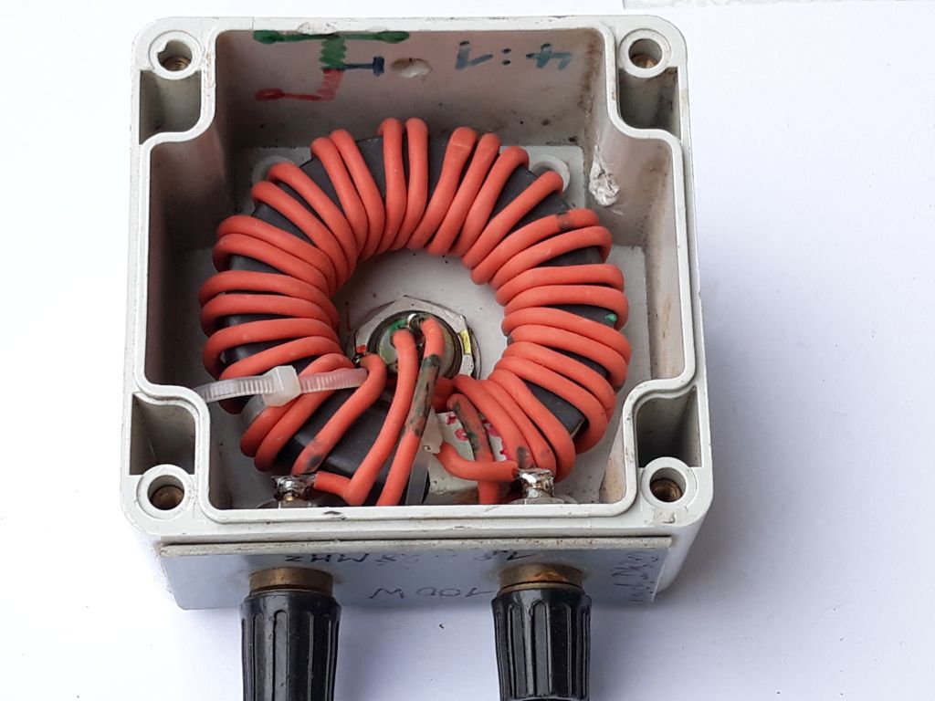 Der folgende Alt-Text wurde noch nicht geprüft: Kurzfassung: Offenes, graues Kunststoffgehäuse mit einem orange umwickelten ringförmigen Kern und zwei schwarzen Anschlüssen an der Unterseite.

Detaillierte Beschreibung: Das quadratische Gehäuse ist von oben auf weißem Hintergrund fotografiert und zeigt innen einen großen, dunkelgrauen Ringkern, der eng mit einem orangefarbenen Draht spiralförmig bewickelt ist. Der Ringkern ist mittig mit einer Schraube und Mutter fixiert; die Drahtenden führen zur Mitte und sind an dortigen Lötpunkten befestigt, ein kleiner Kabelbinder hält eine Drahtschlaufe. Links und rechts im Gehäuseinneren sind Metallschrauben mit Lötösen zu sehen, außerdem Kleber- oder Lötspuren sowie leichte Verschmutzungen und Kratzer. An den Innenwänden sind handschriftliche Markierungen in Grün und Blau, darunter „1:7“ und eine kleine Skizze. Die Ecken haben Schraublöcher mit Messinggewinden. Unten am Gehäuse ragen zwei gerändelte, schwarze Anschlüsse nach außen.