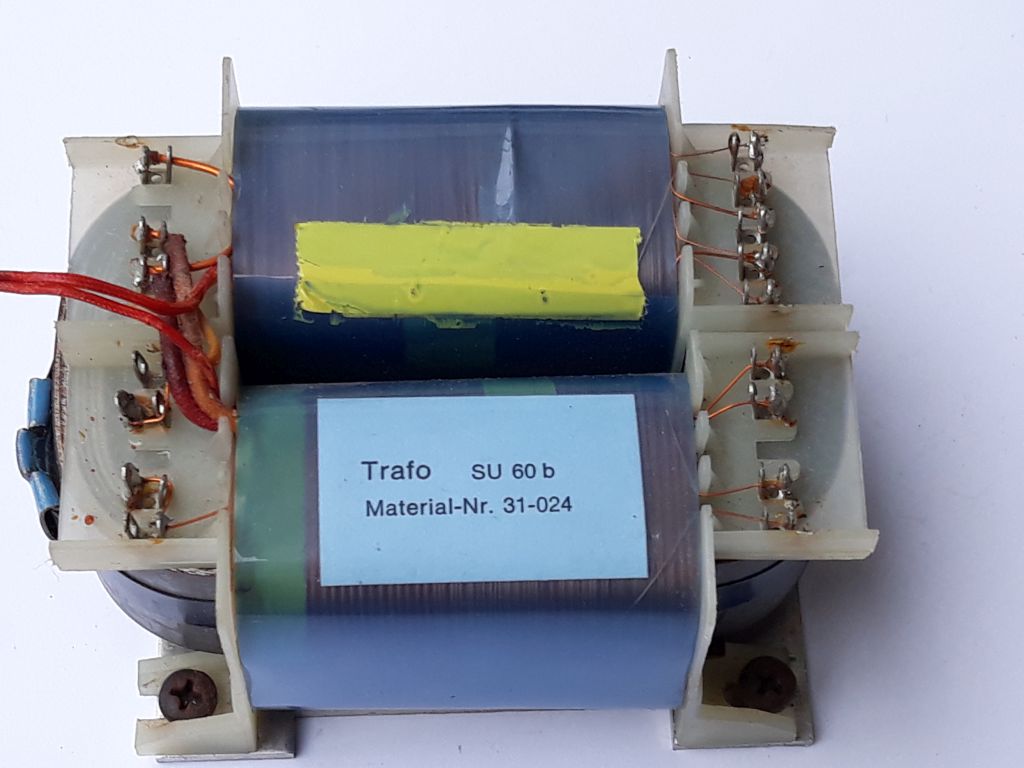 Der folgende Alt-Text wurde noch nicht geprüft: Kurzbeschreibung: Ein Transformator mit zwei Wicklungen und seitlichen Anschlussleisten, von oben auf weißem Hintergrund fotografiert.

Detaillierte Beschreibung: Das Gerät besteht aus einem rechteckigen Blechkern mit zwei nebeneinander liegenden Spulenkörpern, die mit blaugrünem Isoliermaterial umwickelt sind. Auf der oberen Spule klebt mittig ein gelber Streifen, auf der unteren Spule ein hellblaues Etikett mit der Aufschrift „Trafo  SU 60 b“ und „Material-Nr. 31-024“. An beiden Stirnseiten befinden sich helle Kunststoffträger mit Reihen von metallischen Lötfahnen; daran sind feine, lackierte Kupferdrähte angeschlossen, teils mit sichtbaren Lötstellen und leichten Anlaufspuren. Links führen zwei rote flexible Leitungen zu einer Anschlussseite; daneben ist ein kleines blaues zylindrisches Element zu sehen. Unten sind Montagewinkel mit Schrauben erkennbar. Der Hintergrund ist einheitlich weiß, das Foto zeigt das Bauteil leicht von oben.