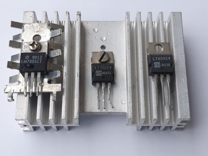 Dieser Alt-Text wurde noch nicht überprüft.

Zusammenfassung: Drei Halbleiterbauteile im TO-220-Gehäuse sind auf bzw. vor silbernen Aluminium-Kühlkörpern angeordnet und von oben auf hellem Hintergrund fotografiert.

Detaillierte Beschreibung: Links sitzt ein schwarzes Bauteil mit Metalllasche und der sichtbaren Aufschrift „LM7905CT 9012“ mit einer Schraube an einem gerippten Kühlkörper; seine drei Anschlussbeinchen zeigen nach unten. In der Mitte steht ein „L7812CV“ (zusätzliche Markierung „SSS 86476“) mit leicht gebogenen Beinchen vor einer flachen Aluminiumplatte mit zwei kleinen Wassertropfen und einem Senkloch; in der Lasche steckt eine kleine Schraube. Rechts ist ein weiteres Bauteil „L7809CV“ (Markierung „SSS 8613S“) an einem zweiten gerippten Kühlkörper befestigt. Alle drei Bauteile haben eine Metalllasche mit Befestigungsloch, die Kühlkörper sind silbern und längs gerippt, und der Hintergrund ist weiß.