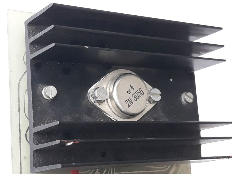 Dieser Alt-Text wurde noch nicht überprüft.

1. Kurze Zusammenfassung: Nahaufnahme eines schwarzen Kühlkörpers mit mehreren Lamellen und einem zentral montierten Metallbauteil mit der Aufschrift „2N3055“.

2. Detaillierte Beschreibung: Im Bild dominiert ein schwarzer Aluminium-Kühlkörper mit vier horizontalen Kühlrippen, montiert auf einer schwarzen Trägerplatte. In der Mitte sitzt ein silbernes, rundes Metallgehäuse mit ovaler Grundplatte, das mit zwei Schrauben befestigt ist; darauf sind „C5“ und „2N3055“ lesbar. Links und rechts in der Trägerplatte befinden sich zusätzlich zwei weitere Schlitzschrauben. Am Rand des Metallgehäuses sind helle, unregelmäßige Rückstände sichtbar. Unten ragt der Rand einer Leiterplatte mit feinen Leiterbahnen ins Bild; am unteren linken Bereich ist ein kurzes Stück einer rot isolierten Leitung zu sehen. Der Hintergrund ist weiß, die Beleuchtung erzeugt leichte Reflexe auf Metall und Lackflächen.