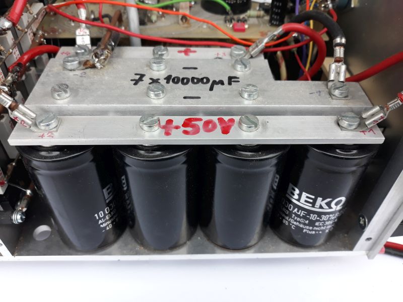Der folgende Alt-Text wurde noch nicht geprüft: 1. Kurze Zusammenfassung:
Vier große, schwarze Elektrolytkondensatoren sind in einem Metallgehäuse montiert und über zwei verschraubte Aluminiumschienen mit roten Anschlusskabeln verbunden, mit handschriftlichen Plus/Minus-Markierungen und der Aufschrift „+50V“ sowie „7 × 10000 µF“.

2. Detaillierte Beschreibung:
Im Vordergrund befinden sich vier zylinderförmige, schwarze Kondensatoren in einer Reihe, mit hellen Aufdrucken wie „10000 µF -10…+30%“ und dem Markenaufdruck „BEKO“; sie stehen in einer Metallwanne. Darüber liegen zwei parallel verlaufende, silberfarbene Aluminiumschienen, die mit mehreren Sechskantschrauben und Unterlegscheiben verschraubt sind. Auf der oberen Schiene sind handschriftliche Kennzeichnungen zu sehen: ein rotes „+“, ein schwarzes „-“, sowie in schwarzer Schrift „7 × 10000 µF“. Auf der unteren Schiene steht in roter Schrift „+50V“. An den Enden und an mehreren Punkten der Schienen sind rote Leitungen mit Ringkabelschuhen angeschlossen; einige Anschlüsse sind mit transparenten Isolierschläuchen versehen. Im Hintergrund sind weitere Bauteile, Leiter, Lötstellen und mehrfarbige Kabel innerhalb des Metallchassis sichtbar. Die Szene zeigt eine geordnete, verschraubte Verdrahtung, nummerierte Markierungen an einzelnen Schrauben und ein insgesamt sauber aufgebautes Modul innerhalb eines größeren elektronischen Geräts.