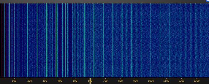 Der folgende Alt-Text wurde noch nicht geprüft: Zusammenfassung: Blaue Spektrums-/Wasserfallanzeige mit vielen schmalen, senkrechten hellen Streifen und einer gelb markierten Skala am unteren Rand.

Detaillierte Beschreibung: Das Bild zeigt eine rechteckige, farbcodierte Darstellung in überwiegend dunklen Blautönen mit körniger Textur. Über die gesamte Höhe verlaufen zahlreiche schmale, helle vertikale Linien in Cyan bis Türkis; sie sind links und im mittleren Bereich dichter und variieren in Helligkeit und Breite. Rechts sind die Linien insgesamt schwächer und teilweise regelmäßiger verteilt. Über die Breite liegen zusätzlich gleichmäßig verteilte, sehr feine vertikale Hilfslinien. Am äußersten linken Rand ist ein schmaler, dunkler Rot- bis Purpurstreifen sichtbar. Unten verläuft ein dunkler Balken mit gelben Markierungen und Zahlen von 0 bis 1300 (Einheit nicht angegeben); eine vertikale grüne Markierungslinie steht ungefähr bei der 600, weitere grüne Linien sind über die Breite verteilt. Insgesamt wechselt die Helligkeit in den blauen Flächen leicht, wodurch ein unruhiges, rauschähnliches Muster entsteht.