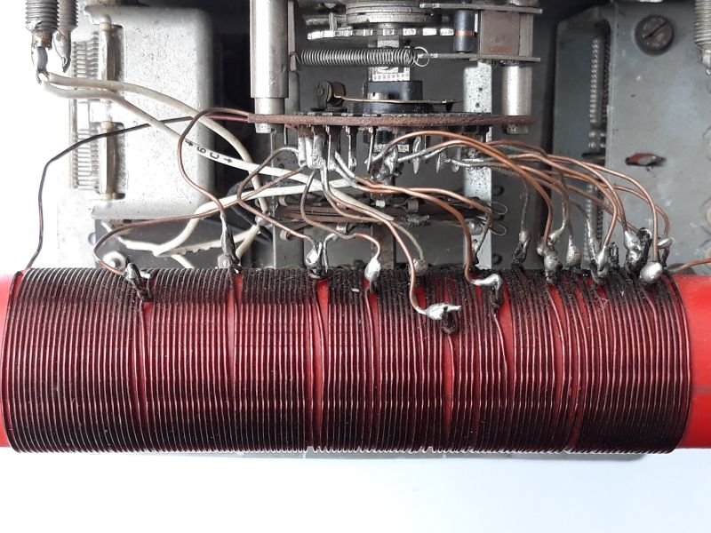 Der folgende Alt-Text wurde noch nicht geprüft: Zusammenfassung: Nahaufnahme des Innenraums eines elektronischen Geräts mit einer dicht gewickelten roten Zylinderspule und zahlreichen Drahtabgriffen, die zu einem mehrstufigen Drehschalter im Metallchassis führen.

Detaillierte Beschreibung: Im unteren Bildbereich liegt horizontal eine rote, zylindrische Spule, eng mit dunkelrotem Kupferdraht bewickelt. Entlang der oberen Spulenseite sind viele Abgriffe sichtbar: dünne, bräunliche lackierte Drähte, die an einzelnen Windungen angelötet sind; an den Lötstellen sitzen silbrig glänzende Lottropfen, teils mit dunklen Verfärbungen an der Umgebung. Die Abgriffe verlaufen bogenförmig nach oben und vorne zu einem gestapelten, kreisförmigen Drehschalter mit mehreren Isolatorscheiben und radial angeordneten Lötfahnen. In der Schaltermitte sitzt eine Metallwelle mit Federn und kleinen mechanischen Teilen. Links und rechts erkennt man das graue Metallchassis mit Schrauben, Federn und Halterungen; einzelne weiße und graue Leitungen führen in den Aufbau. Der Hintergrund ist überwiegend grau-metallisch, unter der Spule ist ein heller, weißer Untergrund zu sehen. Der Gesamteindruck ist technisch, gebraucht und funktional, mit leicht unordentlich geführten Drähten.
