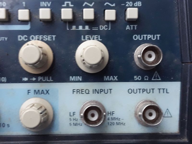 Der folgende Alt-Text wurde noch nicht geprüft: Zusammenfassung: Ausschnitt der Frontplatte eines elektronischen Messgeräts mit mehreren Drehknöpfen und BNC-Buchsen für Ausgang und Frequenzeingang.

Detaillierte Beschreibung: Oben verläuft eine Reihe hellgrauer, quadratischer Tasten mit Symbolen (u. a. Wellenformen) und Beschriftungen wie INV, DC und -20 dB (ATT). Darunter links ein großer, beigefarbener Drehknopf mit der Beschriftung DC OFFSET und dem Hinweis → PULL; in der Mitte ein gleich großer Knopf LEVEL mit Skala von MIN bis MAX. Rechts daneben eine silberne, runde BNC-Buchse mit der Beschriftung OUTPUT und 50 Ω sowie einem Warnsymbol. Im unteren, hellblauen Bereich befindet sich links ein weiterer Drehknopf F MAX. In der Mitte ist eine BNC-Buchse unter der Überschrift FREQ INPUT, umgeben von den Angaben LF 5 Hz–5 MHz und HF 4 MHz–120 MHz. Rechts unten eine weitere BNC-Buchse mit der Beschriftung OUTPUT TTL und einem Warnsymbol. Die Front ist dunkelblau mit hellblauen Feldern; die Bedienelemente wirken leicht benutzt.