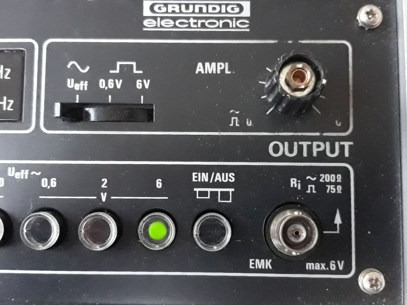 Der folgende Alt-Text wurde noch nicht geprüft: Zusammenfassung: Ausschnitt der Frontplatte eines schwarzen elektronischen Geräts mit der Aufschrift „GRUNDIG electronic“, Reglern, beleuchteter Taste und BNC-Anschluss, beschriftet mit „OUTPUT“.

Detaillierte Beschreibung: Oben mittig steht das Logo „GRUNDIG electronic“. Darunter links befindet sich ein horizontaler Schiebeschalter unter Symbolen für Sinus und Rechteck sowie der Beschriftung „Ueff 0,6V 6V“. Rechts davon ist ein gerändelter Drehknopf mit der Beschriftung „AMPL.“. Auf Höhe dieser Elemente steht groß „OUTPUT“. In der unteren Reihe sind vier runde Drucktasten/Linsen angeordnet; die dritte von links leuchtet grün. Darüber steht „Ueff ~ 0,6 2 6 V“, rechts daneben „EIN/AUS“ mit einem zweistufigen Symbol. Rechts unten sitzt eine runde Metallbuchse (BNC) mit Pfeilsymbol, daneben die Beschriftungen „Ri ~ 200Ω 75Ω“ und „EMK max. 6 V“. Die Bereiche sind durch weiße Linien eingerahmt; die Front ist schwarz mit weißer Beschriftung und hat sichtbare Schrauben in den Ecken.