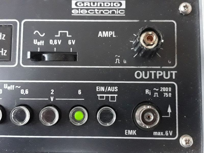 Dieser Alt-Text wurde noch nicht überprüft.

Zusammenfassung: Ausschnitt der Frontplatte eines schwarzen elektronischen Geräts mit der Aufschrift „GRUNDIG electronic“, Reglern, beleuchteter Taste und BNC-Anschluss, beschriftet mit „OUTPUT“.

Detaillierte Beschreibung: Oben mittig steht das Logo „GRUNDIG electronic“. Darunter links befindet sich ein horizontaler Schiebeschalter unter Symbolen für Sinus und Rechteck sowie der Beschriftung „Ueff 0,6V 6V“. Rechts davon ist ein gerändelter Drehknopf mit der Beschriftung „AMPL.“. Auf Höhe dieser Elemente steht groß „OUTPUT“. In der unteren Reihe sind vier runde Drucktasten/Linsen angeordnet; die dritte von links leuchtet grün. Darüber steht „Ueff ~ 0,6 2 6 V“, rechts daneben „EIN/AUS“ mit einem zweistufigen Symbol. Rechts unten sitzt eine runde Metallbuchse (BNC) mit Pfeilsymbol, daneben die Beschriftungen „Ri ~ 200Ω 75Ω“ und „EMK max. 6 V“. Die Bereiche sind durch weiße Linien eingerahmt; die Front ist schwarz mit weißer Beschriftung und hat sichtbare Schrauben in den Ecken.