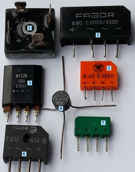 Der folgende Alt-Text wurde noch nicht geprüft: Kurzfassung: Auf weißem Hintergrund liegen mehrere verschiedenfarbige elektronische Bauteile mit Anschlussdrähten und aufgedruckten Symbolen (+, −, ~), teils mit blauen Nummernkästchen (1, 2, 4, 5, 6, 7) markiert.

Details:
- Oben links (1): Schwarzer, annähernd quadratischer Block mit zentraler runder Metallbuchse; Aufdruck „AC“, sowie „+“ und „−“ auf der Vorderseite.
- Oben rechts (2): Schwarzes, rechteckiges Gehäuse mit vier nach unten zeigenden Pins; Aufdruck „FAGOR B80 C5000/3300“ und Symbole „~“, „+“, „−“.
- Mitte links (ohne Nummer): Schwarzes, rechteckiges Bauteil mit vier Pins; Aufdruck „BY225 100 E7033“ sowie Symbole „~“, „+“, „−“.
- Bildmitte (4): Kleines rundes, schwarzes Bauteil (Scheibenform) mit vier langen, kreuzförmig abstehenden Drähten; Aufdruck u. a. „GIE C1000C“.
- Rechts daneben (5): Orangefarbenes, rechteckiges Gehäuse mit vier kurzen Pins; Aufdruck mit Logo und „B 40 C 1500“ sowie Polaritäts- und Wechselspannungssymbole.
- Unten links (6): Schwarzes, trapezförmiges Gehäuse mit vier Pins; Aufdruck „FAGOR FBU 4M“ und Symbole für „+“ und „−“.
- Unten rechts (7): Grünes, rechteckiges Bauteil mit vier Pins; kleine runde Vertiefungen oben und Aufdruck mit „+“, „−“ und „~“-Symbolen.
- Zusätzlich: Ein einzelner, langer, dünner Draht verläuft im Zentrum vertikal nach oben.
