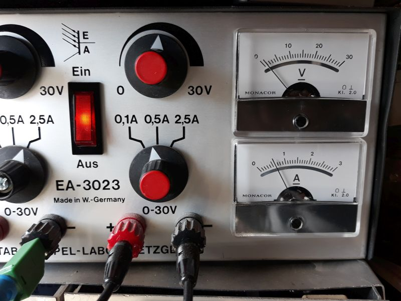 Dieser Alt-Text wurde noch nicht überprüft.

1. Zusammenfassung: Nahaufnahme der Frontplatte eines Geräts EA‑3023 mit zwei analogen Anzeigen (Volt und Ampere), zwei großen Drehknöpfen, einem rot leuchtenden Wippschalter und mehreren farbigen Anschlussbuchsen mit eingesteckten Leitungen.

2. Detaillierte Beschreibung: Links sind zwei schwarze Drehknöpfe mit roten Mitten zu sehen; um den oberen Knopf ist „0 … 30V“ markiert, um den unteren „0,1A 0,5A 2,5A“. Dazwischen befindet sich ein rechteckiger, rot beleuchteter Wippschalter mit der Beschriftung „Ein“ oben und „Aus“ unten. Darüber steht ein stilisiertes „EA“-Logo, daneben mehrfach „30V“. Im unteren linken Bereich steht „EA‑3023“ und „Made in W.-Germany“. Entlang der unteren Kante sind mehrere 4‑mm‑Buchsen und Stecker sichtbar: u. a. ein grüner, ein roter und ein schwarzer Stecker mit angeschlossenen Kabeln; neben den Buchsen ist „0‑30V“ aufgedruckt. Rechts sind zwei übereinander angeordnete analoge Messwerke der Marke „MONACOR“: oben mit der Skala „0–30“ und dem Symbol „V“ (Volt), unten mit der Skala „0–3“ und dem Symbol „A“ (Ampere); beide Instrumente tragen die Kennzeichnung „Kl. 2.0“. Das Gehäuse wirkt metallisch/silbern, die Beschriftungen sind schwarz, Bedienelemente überwiegend schwarz mit roten Akzenten.