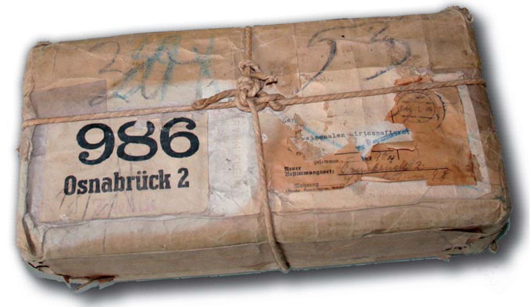 Der folgende Alt-Text wurde noch nicht geprüft: Zusammenfassung: Ein rechteckiges, braun eingepacktes Paket mit Schnurverschnürung und stark abgenutzten Etiketten, auf denen groß „986 Osnabrück 2“ steht.

Detaillierte Beschreibung: Das Paket ist in braunes, zerknittertes und stellenweise eingerissenes Packpapier gewickelt, das mit mehreren Klebestreifen geflickt wurde. Eine grobe, helle Kordel verläuft längs und quer über die Oberfläche und ist oben mittig zu einem Knoten gebunden. Links befindet sich ein weißes, verschmutztes Label mit der schwarzen, großen Zahl „986“ und darunter „Osnabrück 2“. Rechts klebt ein weiteres, stark verwittertes Adress- oder Formularetikett mit verblasster Schreibmaschinen- und Handschrift, partiellen Papierabrissen und einem runden Stempelabdruck oben rechts. Über das Papier ziehen sich blaue Wachskreide- bzw. Bleistiftmarkierungen, darunter große Ziffern und Pfeilformen. Die Ecken sind eingedrückt und ausgefranst; das Paket liegt leicht schräg und wirft einen weichen Schatten auf den hellen Hintergrund.