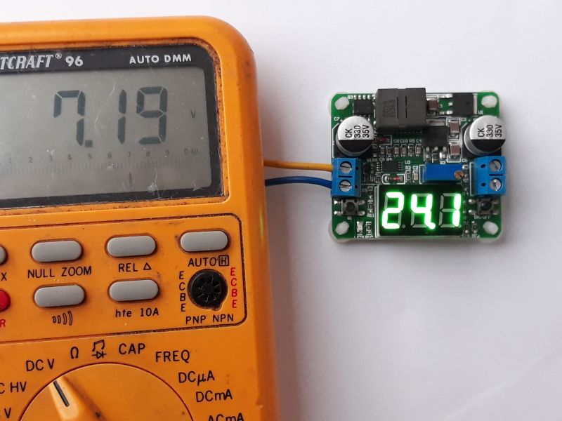 Der folgende Alt-Text wurde noch nicht geprüft: Kurzbeschreibung: Ein oranges Digitalmultimeter liegt neben einer kleinen Elektronik‑Platine mit grünem LED‑Display; das Multimeter zeigt 7.19 V, die Platine 24.1.

Detaillierte Beschreibung: Links dominiert ein abgenutztes, orangefarbenes Hand‑Digitalmultimeter mit großem grauen LCD, auf dem „7.19“ steht, rechts daneben ein kleines „V“. Darunter sind mehrere graue Gummitasten mit Aufdrucken wie „MAX“, „REL“ und „AUTO“, ein schwarzer runder hFE‑Sockel sowie ein Drehschalter mit Skalen für DCV, Ω, CAP und FREQ. Rechts daneben liegt eine kleine grüne Leiterplatte mit bestückten Bauteilen: zwei silberne Elektrolytkondensatoren mit „35V“-Aufdruck, ein schwarzer rechteckiger Induktor, mehrere ICs und Widerstände sowie zwei blaue Schraubklemmen. Auf der Platine leuchtet eine dreistellige grüne 7‑Segment‑Anzeige mit „24.1“. Ein gelbes und ein blaues Kabel sind an eine der Klemmen angeschlossen und führen nach links aus dem Bild. Der Hintergrund ist eine weiße, matte Fläche.