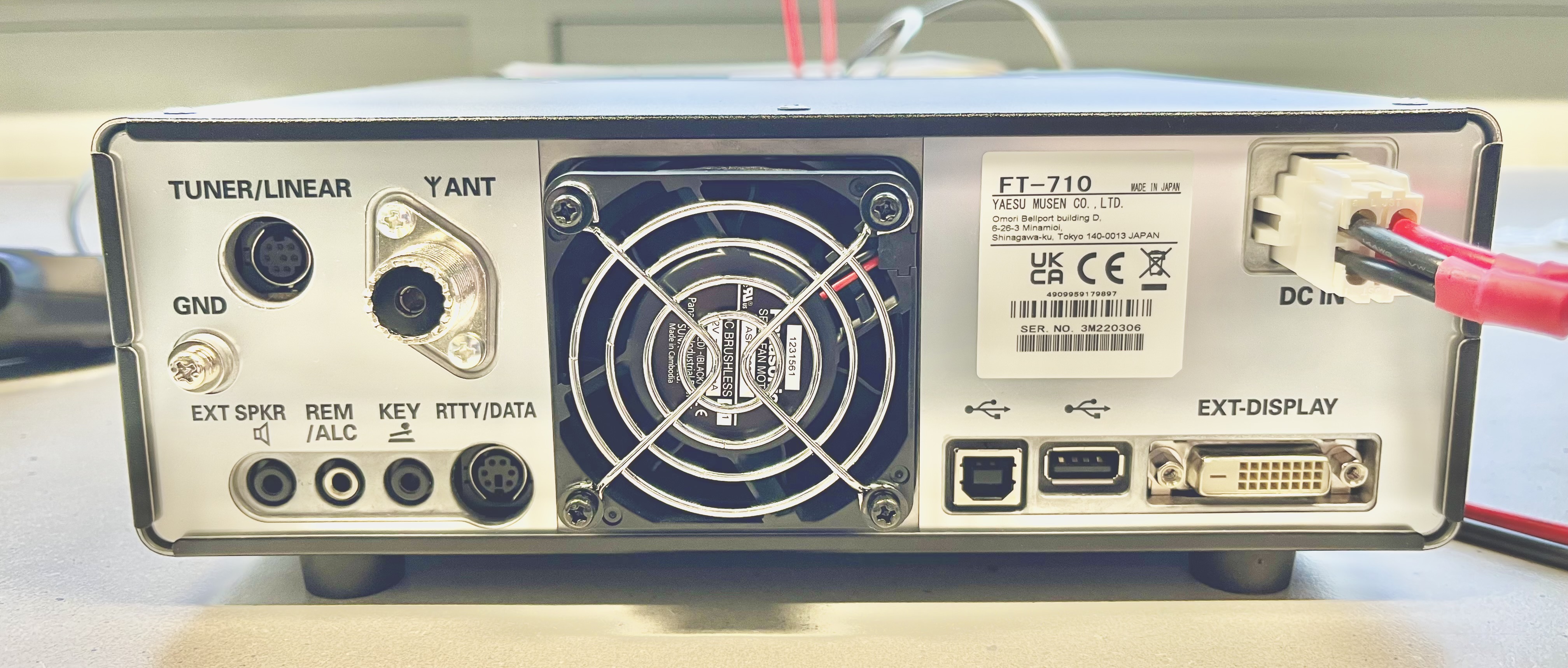 Der folgende Alt-Text wurde noch nicht geprüft: 1. Kurze Zusammenfassung: Rückseite eines elektronischen Geräts (Modell FT‑710) mit vielen Anschlüssen, zentralem Lüftergitter und angeschlossenem DC‑Stromkabel.

2. Detaillierte Beschreibung: Das Gerät steht auf einem hellen Tisch; im Zentrum befindet sich ein großer, runder, mit vier Schrauben befestigter Lüfter mit Metall-Schutzgitter und sichtbarem „Brushless Fan“-Aufkleber. Links sind mehrere Buchsen und Beschriftungen: oben „TUNER/LINEAR“ an einer runden mehrpoligen Mini‑DIN‑Buchse, darunter „GND“ an einem Massebolzen mit Rändelmutter, daneben „Y ANT“ bei einer großen koaxialen SO‑239‑Buchse. In der unteren linken Reihe stehen nebeneinander Klinkenbuchsen und Mini‑DIN‑Buchsen, beschriftet mit „EXT SPKR“, „REM/ALC“, „KEY“ und „RTTY/DATA“. Rechts neben dem Lüfter klebt ein weißes Typenschild mit der Aufschrift „FT‑710“, Hersteller‑ und Zulassungszeichen sowie einer Seriennummer. Ganz rechts oben ist „DC IN“ mit einem weißen Steckverbinder zu sehen, in den rote und schwarze Stromkabel eingesteckt sind. Unten rechts ist der Bereich „EXT‑DISPLAY“ mit mehreren Schnittstellen: zwei USB‑Anschlüsse (links ein quadratischer Typ‑B, daneben ein rechteckiger Typ‑A), ein kleiner gewindeter Koax‑Anschluss und ein weißer DVI‑Anschluss mit zwei Befestigungsschrauben. Der Hintergrund ist unscharf und zeigt eine helle Umgebung.